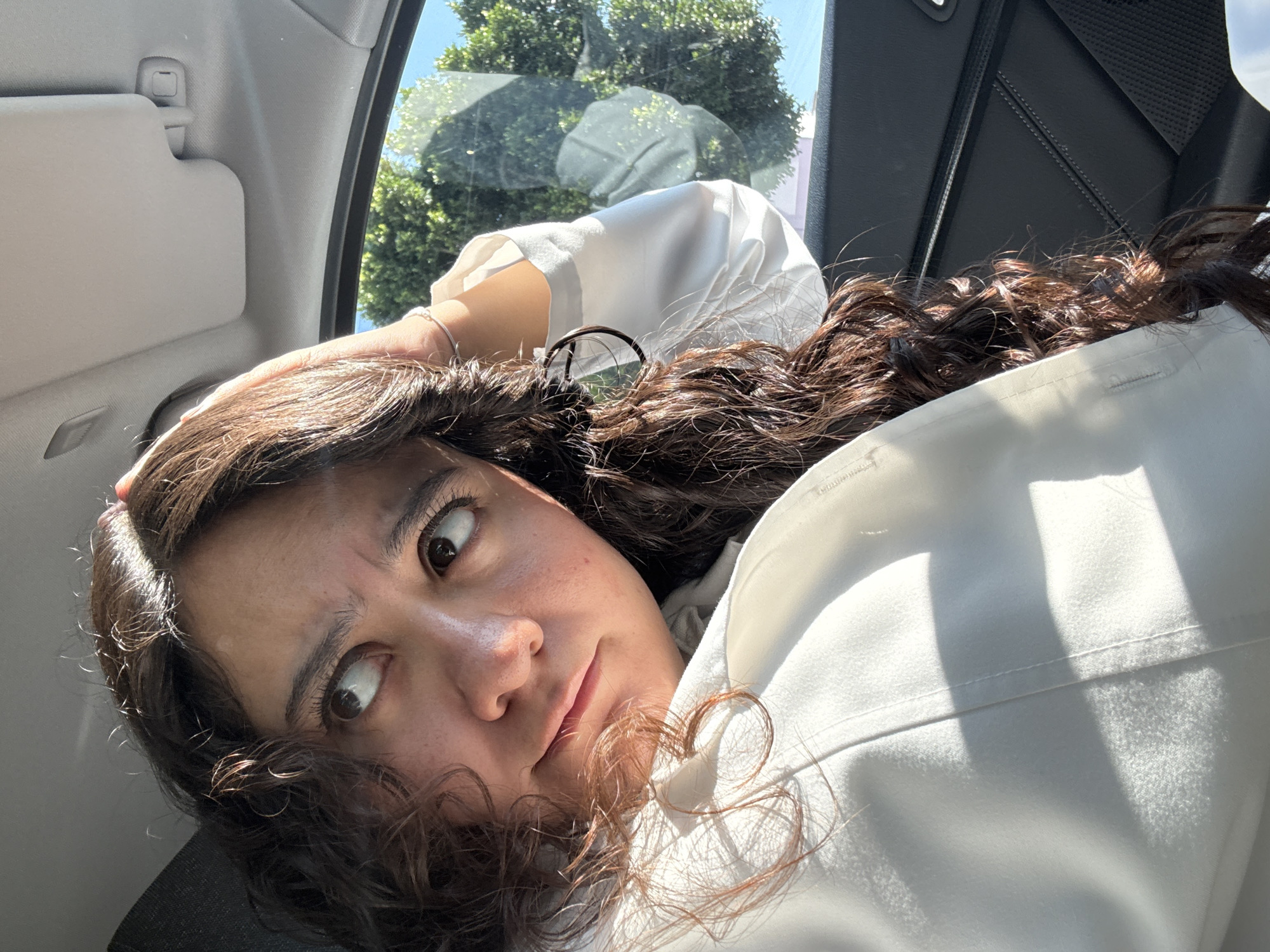A close-up, angled shot captures a young woman inside a car, likely from the back seat. She has dark, curly hair with brown undertones, framing her face, and her head is tilted back, resting against a surface not fully visible. Her wide, dark eyes are fixed on the viewer, accompanied by a slight smirk or playful expression on her lips. Bright sunlight streams in through the car window, illuminating her face and the white long-sleeved shirt she wears, casting sharp shadows on her clothing and hair. A pair of glasses is perched on top of her head, nestled in her hair, and a silver bracelet adorns her left wrist.
Through the car window, the vibrant green foliage of trees and a clear blue sky indicate a sunny outdoor environment. Visible car interior elements include a grey sun visor, a window frame, and part of a seatbelt mechanism. The woman's relaxed posture suggests she is either resting or posing for the photograph. This scene takes place in Puebla, Mexico. There is no visible text.