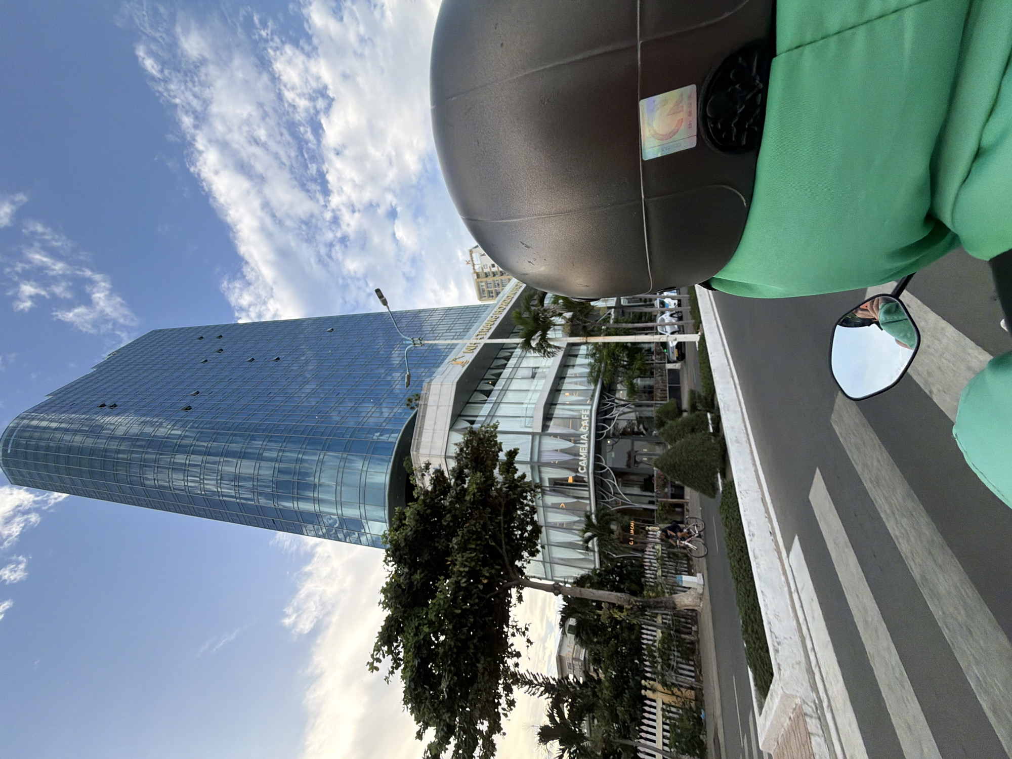 In Nha Trang, Vietnam, a person wearing a green jacket and a dark helmet rides a scooter. The scooter's mirror reflects the rider's face. The scooter is on a paved road, approaching a crosswalk. To the left, a tall, modern building with a blue glass facade dominates the skyline. The building has multiple floors and is partially obscured by a large tree with dark green leaves. Below the glass facade, the building features a white base with the signage "CAMELIA CAFE" visible. Palm trees and other greenery surround the building, and a white picket fence runs along the roadside. In the distance, other buildings and vehicles can be seen. The sky is blue with scattered white clouds.