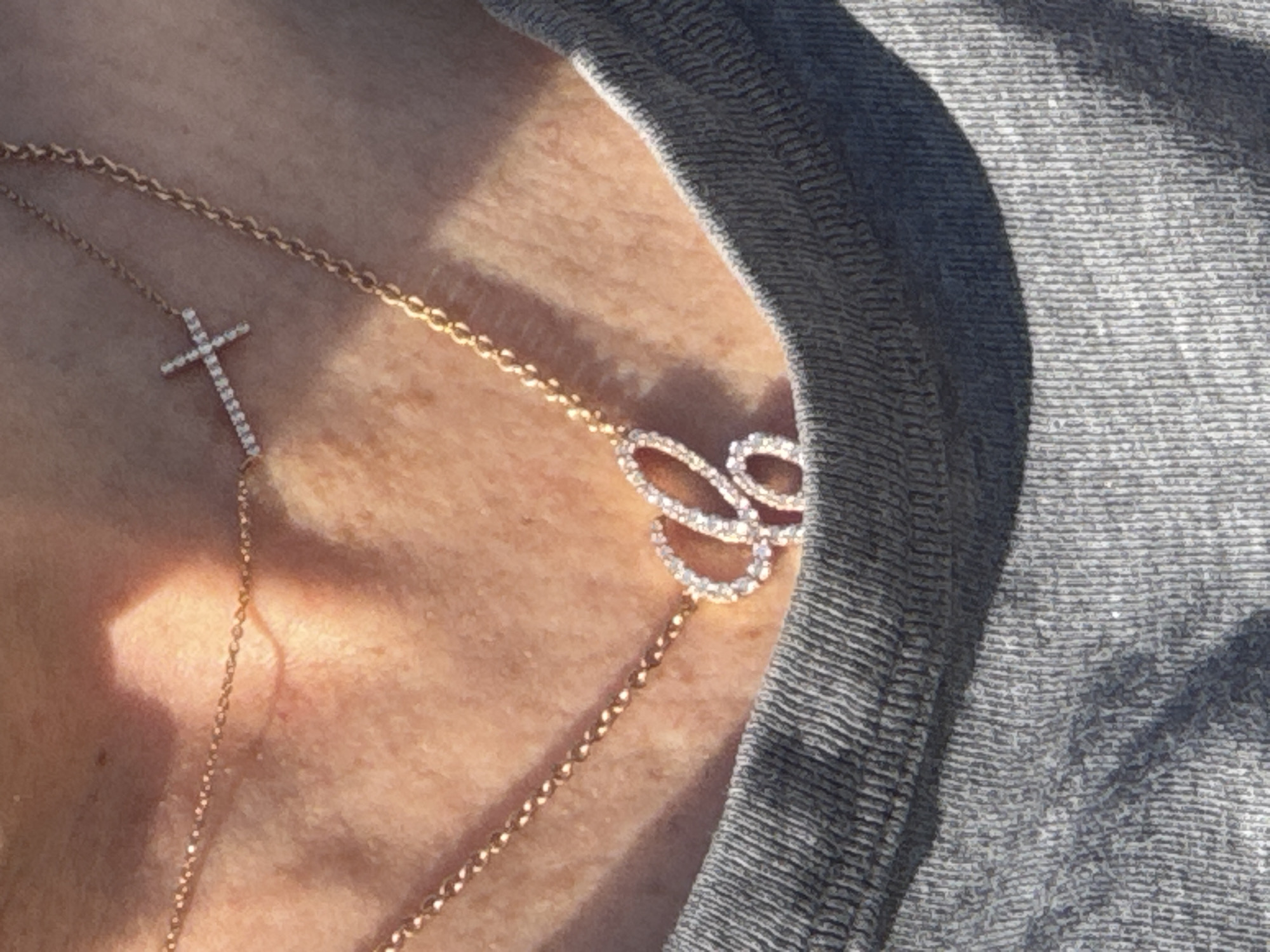 A close-up shot reveals a person wearing two gold necklaces. One necklace features a small cross pendant adorned with sparkling stones, while the other displays a butterfly-shaped charm, also encrusted with stones. The necklaces rest against the person's skin, with a textured gray fabric visible near the right edge of the frame. The setting or location in Vila Nova de Gaia, Portugal is not determinable from this image.
