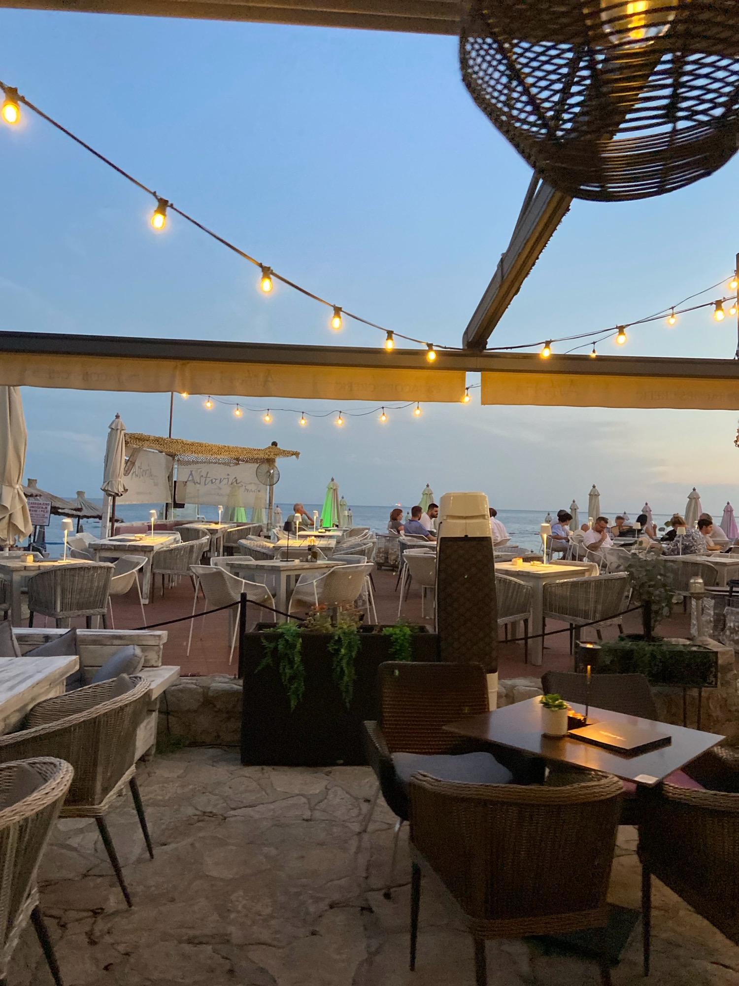 The image shows an outdoor seating area of a restaurant or cafe, likely located on a beachfront in Budva, Montenegro. The time of day appears to be dusk or early evening, as indicated by the twilight sky and the illuminated string lights overhead. Numerous tables with chairs are set up, many of them occupied by diners. The tables are made of wood and have small lamps on them. The chairs are woven wicker. In the background, umbrellas and more tables are visible, suggesting a larger establishment. Beyond the seating area, the sea can be seen stretching to the horizon, with a few boats in the distance. The overall ambiance is relaxed and inviting, with the warm glow of the lights contrasting with the fading daylight. There are several potted plants and some decorative elements, contributing to the atmosphere. The architecture suggests a Mediterranean style, consistent with a coastal town like Budva.