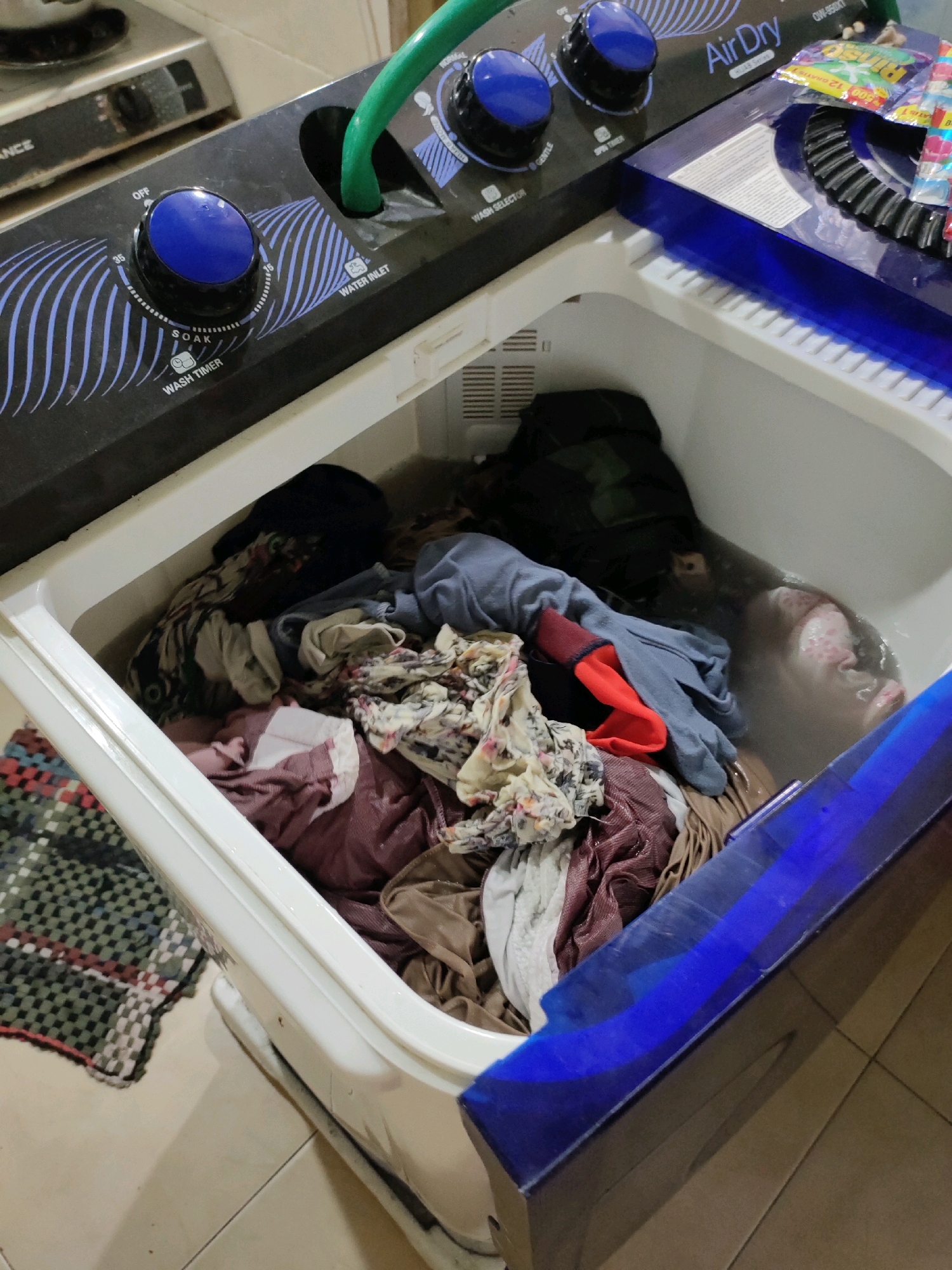 The image shows a top-loading twin tub washing machine filled with water and clothes. The washing machine is white with a blue lid and control panel. The control panel has two blue knobs, one labeled "WASH TIMER" with settings for "SOAK", "OFF", and timer durations like "15", "35", and "55". The other knob is labeled "WASH SELECTOR" with settings like "GENTLE" and "REGULAR". A green hose is connected to the "WATER INLET" on the control panel. The inner tub of the washing machine is partially filled with water and a mix of colorful clothes, including what appears to be a dark blue garment, a patterned shirt, a light blue shirt, and a reddish-brown garment. There is also a small pile of what looks like laundry detergent packets on the lid of the washing machine to the right. The washing machine appears to be situated in a domestic indoor setting, with tiled flooring visible below. The overall scene suggests the process of washing laundry. The location of Kroman, Indonesia, is not explicitly depicted in the image and cannot be determined from the visual information provided.