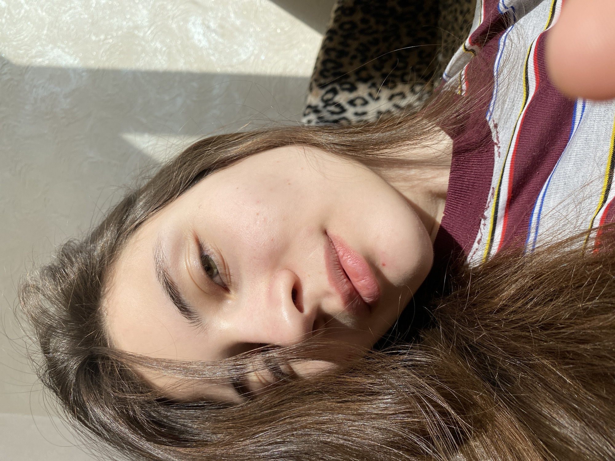 The image presents a close-up portrait of a young person, likely female, with long, dark brown hair. She appears to be lying down, with her head tilted, and her face illuminated by strong, directional sunlight entering from the left. This light casts distinct shadows on a light-colored, textured surface behind her.
Her expression is relaxed and natural. Her right eye, appearing greenish-brown, is open and looks slightly upwards. Her lips are naturally full and slightly parted. Her dark brown hair is spread around her head and face. She is wearing a vertically striped top with colors including maroon, white, light blue, and yellow.
To the right and behind her head, a blurry leopard print pattern is visible, suggesting a pillow, blanket, or furniture. A finger, likely belonging to the photographer or subject, is partially visible in the upper right corner of the original image, hinting at a close-up, possibly a selfie. The strong sunlight indicates it is daytime, likely indoors in a home setting, within the context of Votkinsk, Russia. No visible text is present.