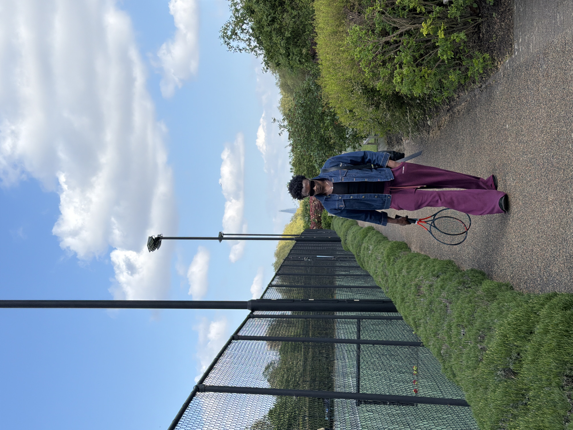 A person stands on a paved path alongside a tennis court in London, United Kingdom, under a clear blue sky with scattered white clouds. The individual, who appears to be male, is positioned towards the center-right of the frame. They have dark skin, short dark hair, and are wearing black sunglasses, a black t-shirt, a dark blue denim jacket, and dark purple sweatpants with unclear white text or a logo on the left leg. A black bag is slung over their shoulder. The person holds a red and black tennis racket in their right hand, pointing downwards.
To the left of the person is a black chain-link fence, beyond which lies a green tennis court. A long, manicured green hedge runs parallel to the fence. Several tall black poles, likely for lighting, are visible above the fence. The paved path continues into the background, and to the person's right, it is bordered by green bushes and trees. In the far distance, a tall, slender building is faintly visible against the sky, partially obscured by foliage. The scene depicts a bright, sunny day in an outdoor recreational setting, likely a park.