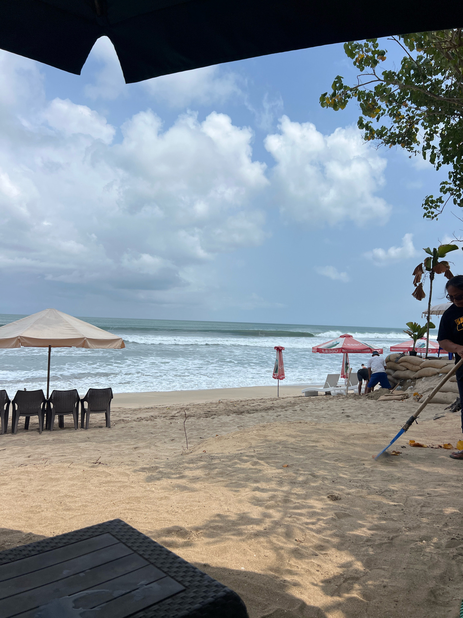 The image shows a beach scene in Kuta, Indonesia. In the foreground, there is a dark table with a woven texture, casting shadows on the sandy beach. Further back, a person is raking the sand with a blue rake. Behind them, several sandbags are piled up, and two other individuals are working on the beach. There are also beach chairs and umbrellas set up, with lounge chairs and red and white beach umbrellas visible in the middle ground. The ocean with waves is in the background, under a cloudy sky. Trees with green and yellow leaves are visible on the right side of the image.