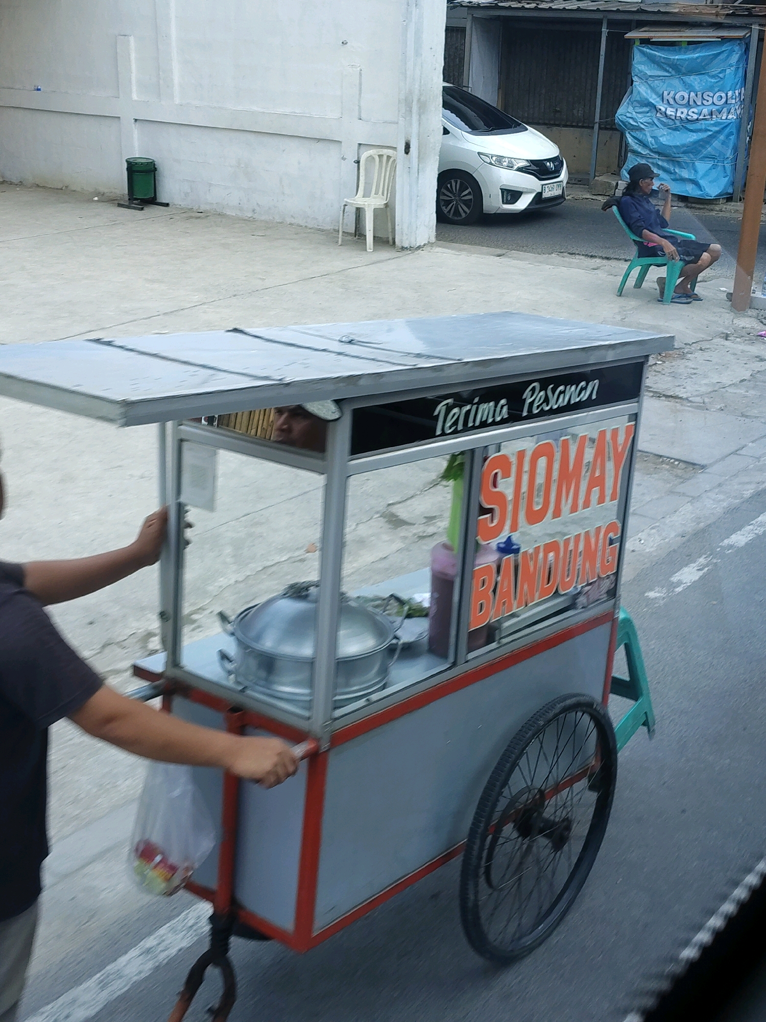 A street vendor is pushing a food cart. The cart has a clear glass display with "TERIMA PESANAN SIOMAY BANDUNG" written on it. Inside the cart, there is a metal pot and a person's face peeking from behind the cart. A plastic bag with some items is hanging from the side of the cart. The cart has a red frame and a silver roof. The street is paved with asphalt, and a white car is parked on the side. In the background, there is a blue tarp with writing on it, and a man is sitting on a green plastic chair. The scene appears to be in a street environment in Tangerang, Indonesia, likely on a sidewalk or near a building.