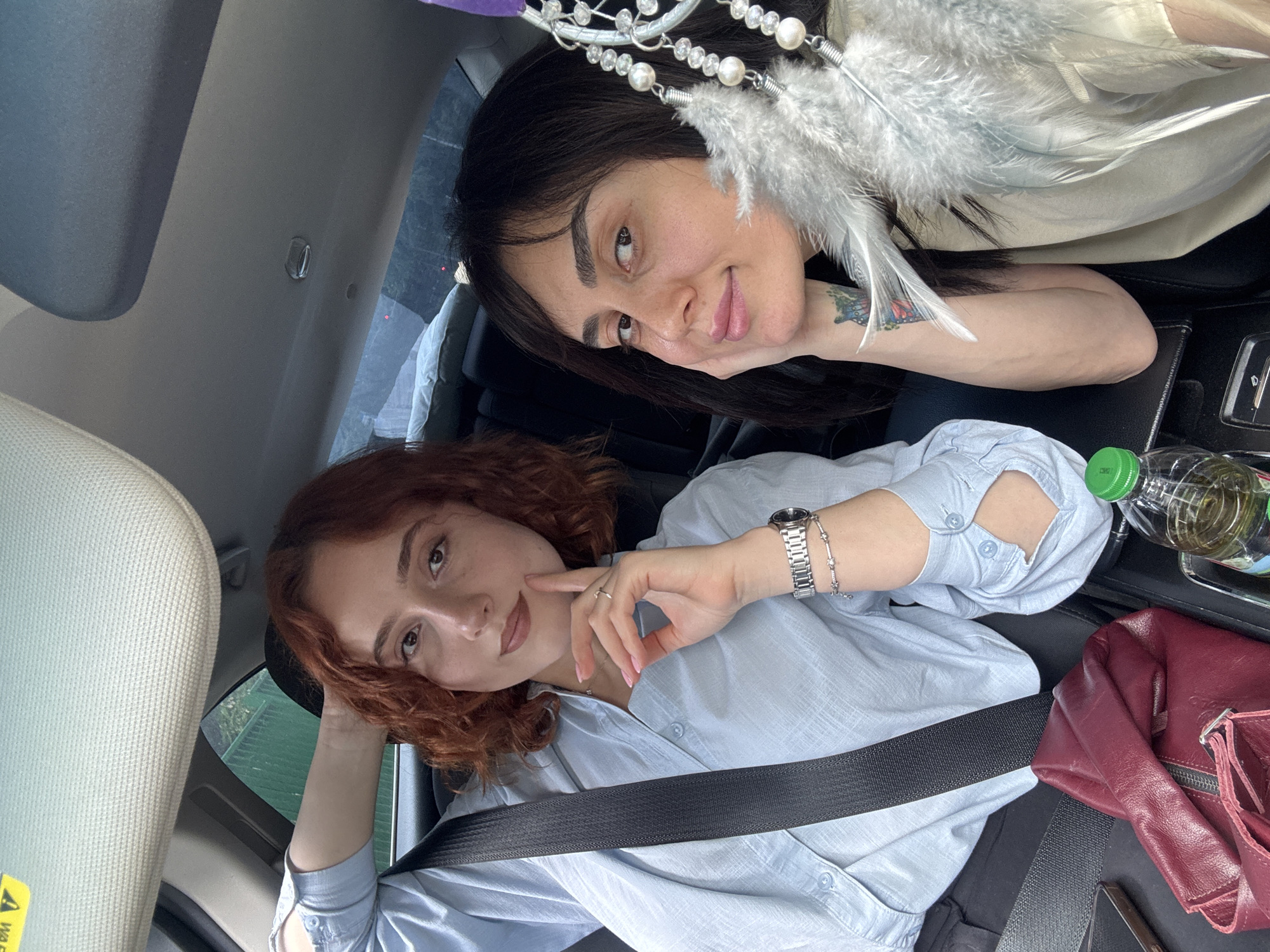 Two women are posing for a selfie inside a car. The woman in the foreground has red curly hair and is wearing a light blue button-down shirt with a seatbelt. She has a watch and bracelet on her left wrist, and a ring on her finger. Her left hand is resting on her cheek. To her right, there is a red bag and a bottle of water. The woman in the background has dark hair and is also wearing a light-colored top. She has a tattoo on her right arm and a dreamcatcher hanging above her. The car's interior is visible, with the sun visor in the foreground and seats in the background. Based on the vehicle registration plate, the image was taken in Tbilisi, Georgia.