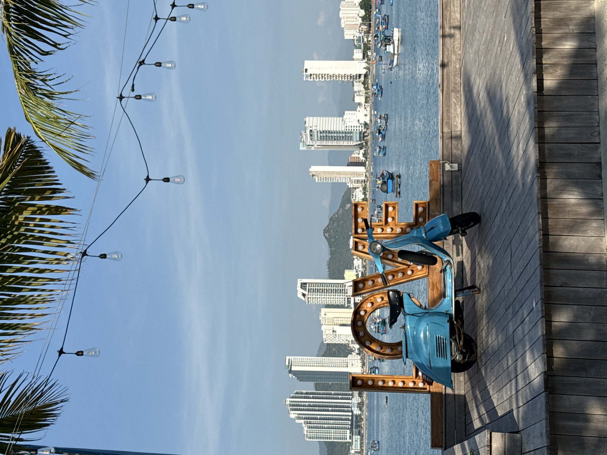 This is a medium shot, eye-level, full-frame photograph taken outdoors on a sunny day. In the foreground, a turquoise blue vintage-style scooter is parked on a wooden deck. Behind the scooter, the word "LOVE" is spelled out in large, illuminated wooden letters, with bulbs embedded around each letter. The "O" letter is partly obscured by the scooter. Beyond the wooden platform, a body of water is visible, filled with numerous small fishing boats. In the background, a city skyline features several tall, modern white buildings, with mountains visible behind them. Palm fronds are seen hanging into the frame from the top edge of the photograph. String lights with exposed bulbs are suspended across the top portion of the image. The scene appears to be a tourist area in Bac Nha Trang Ward, Vietnam, likely a waterfront promenade or park.