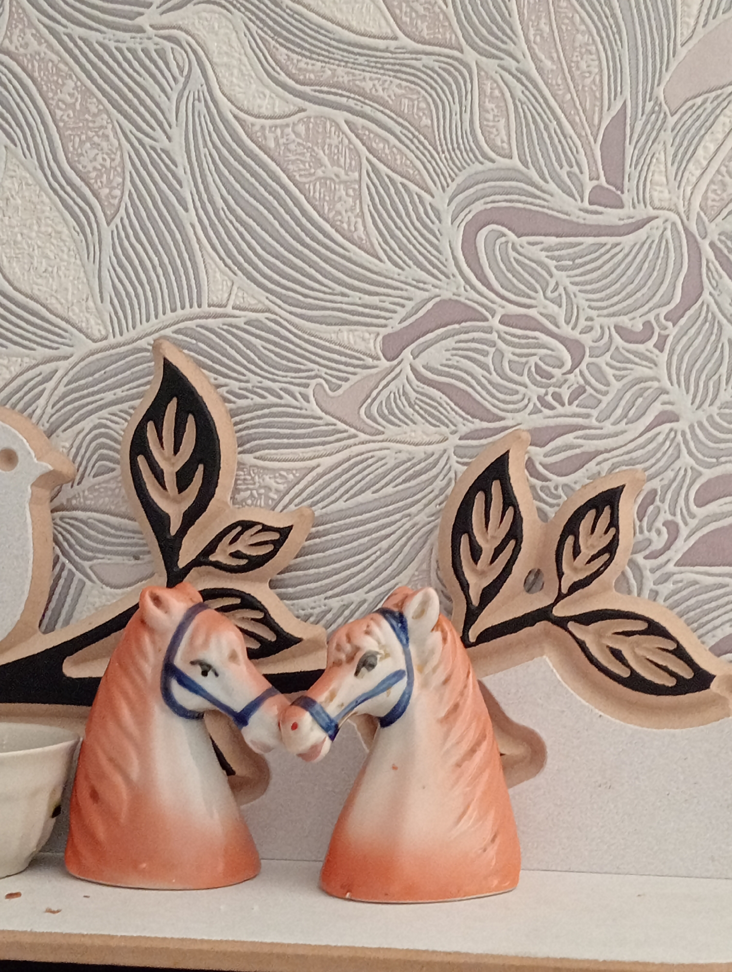 Two ceramic horse figurines are positioned front-on, facing each other, with their noses almost touching. They are a peachy-orange color with white muzzles and black painted eyes, each adorned with a blue halter. The horses are situated on a white shelf in front of a textured wallpaper that features an intricate, abstract pattern of light gray and white lines against a pale pink background. To the left of the horses, a portion of a white ceramic cup is visible. Behind the horses and extending to the left and right is a wooden decorative piece shaped like stylized leaves, painted black and a light wood tone. The lighting appears to be overhead and diffused, creating soft shadows. The scene is still and posed.