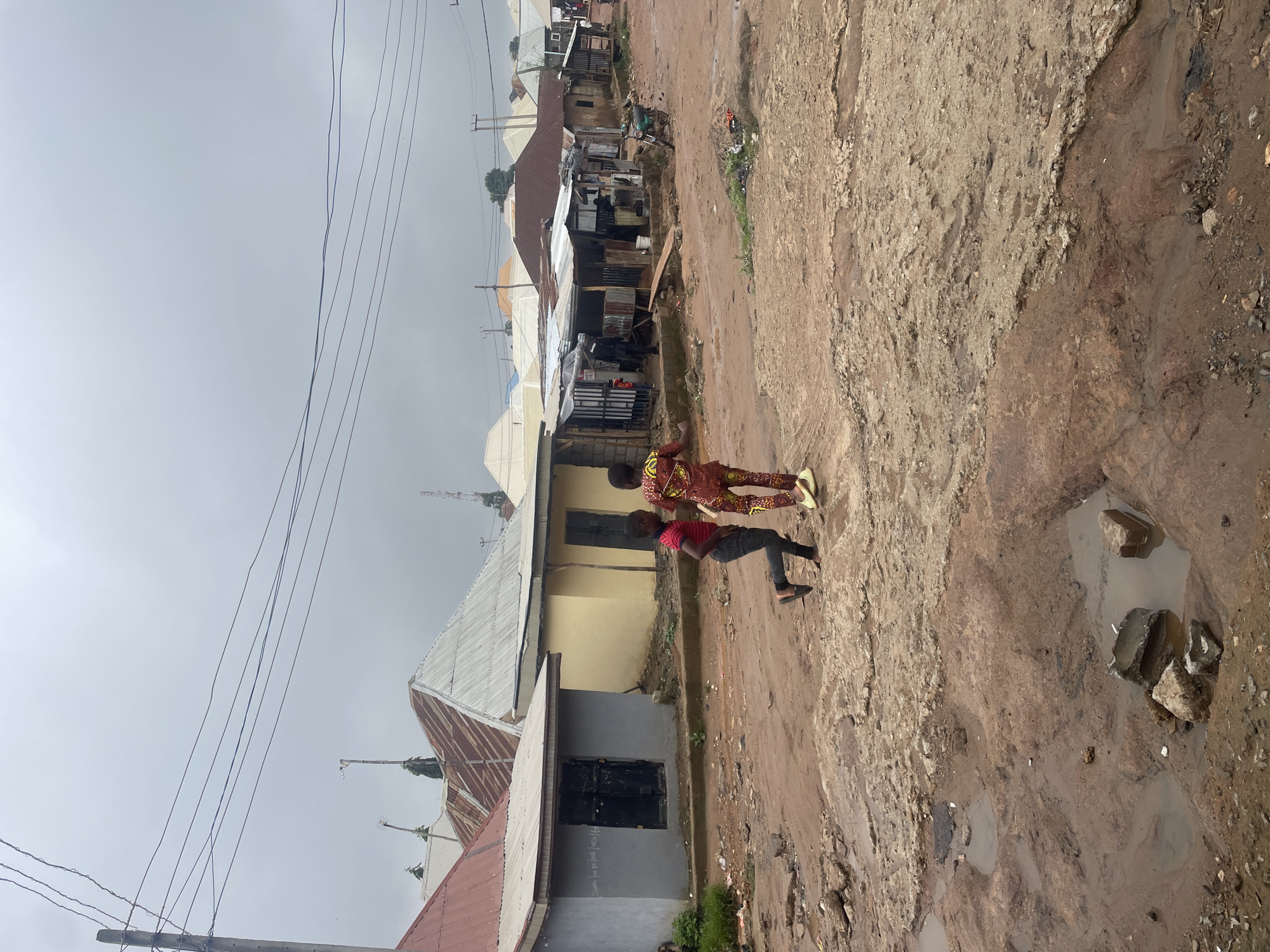 In Kurunduma, Nigeria, two young boys are walking on a muddy, uneven road under an overcast sky. The boy on the left wears a red shirt and dark pants, while the boy on the right is dressed in a vibrant, patterned traditional outfit. They are walking away from the viewer towards a row of modest buildings with corrugated metal roofs. Some buildings appear to be shops or market stalls, with visible goods and structures made of corrugated metal and wood. A motorcycle is parked on the side of the road near one of the shops. The road is littered with debris, including rocks and plastic waste, and contains several large puddles of muddy water, indicating recent rain. Utility poles and power lines are visible in the background, suggesting an inhabited area.