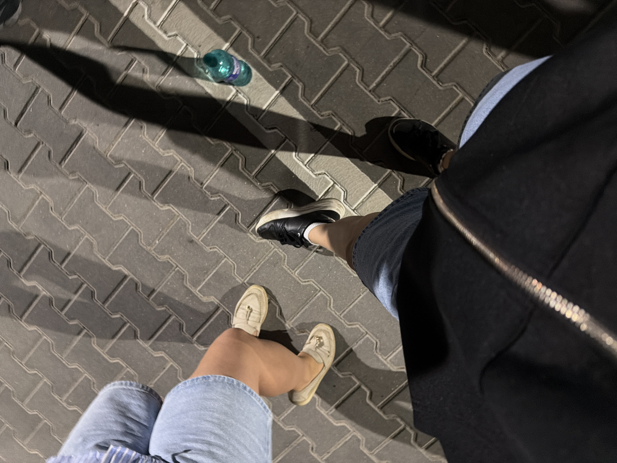 An overhead view captures the legs and feet of two individuals standing on a grey paver stone walkway. The pavers are laid in an interlocking pattern. The person on the left wears light blue denim shorts and light beige slip-on loafers with a tassel detail. The person on the right wears dark blue denim shorts and black sneakers with white soles and laces, and white socks. A partially visible black jacket with a silver zipper runs across the bottom of the frame, obscuring the legs of the person wearing the black sneakers. A partially visible turquoise plastic water bottle stands upright on the walkway to the right of the frame. Long, dark shadows are cast across the pavers, indicating a strong light source, likely the sun. The scene is set in the city of Băcioi, Moldova.