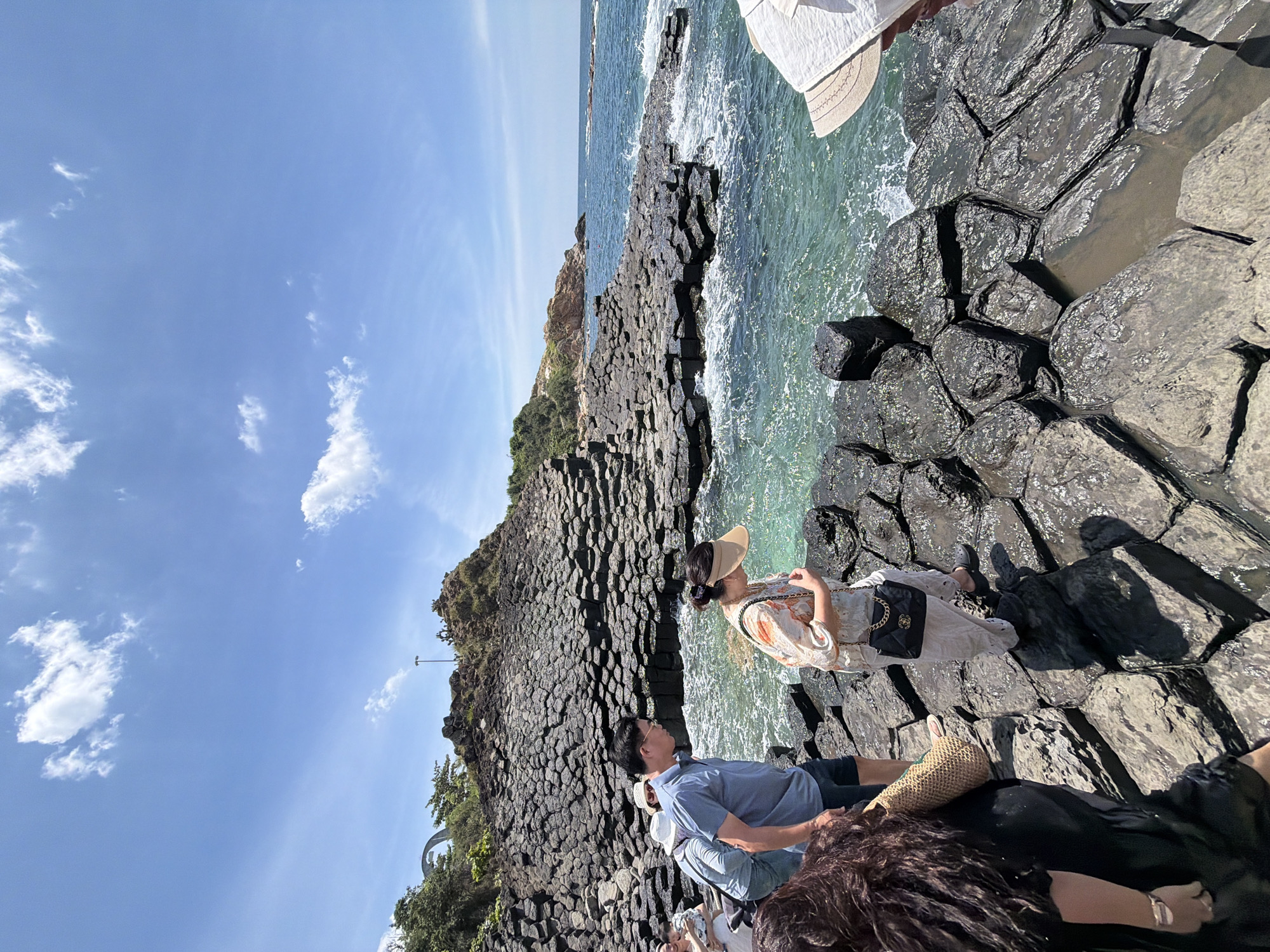 The image depicts a group of people at a seaside location with unique geological formations. The scene appears to be a rocky coastline with hexagonal basalt columns, characteristic of places like Gành Đá Dĩa in Phú Lương, Vietnam.
Several individuals are visible, some are sitting and others are standing. One woman, wearing a sun hat and a patterned dress, is looking out towards the sea. A man in a blue shirt is also present, and other people are partially visible, suggesting a gathering or a shared outing. One person is wearing a white hat. There's a straw bag resting near some of the individuals.
The sea is a vibrant turquoise color, with waves gently breaking against the dark, geometric rocks. The sky is a clear blue with scattered white clouds, indicating a sunny day. The sun appears to be high in the sky, suggesting it's daytime, likely midday or early afternoon. The atmosphere seems relaxed and leisurely, with people enjoying the natural scenery. There is no visible text in the image.