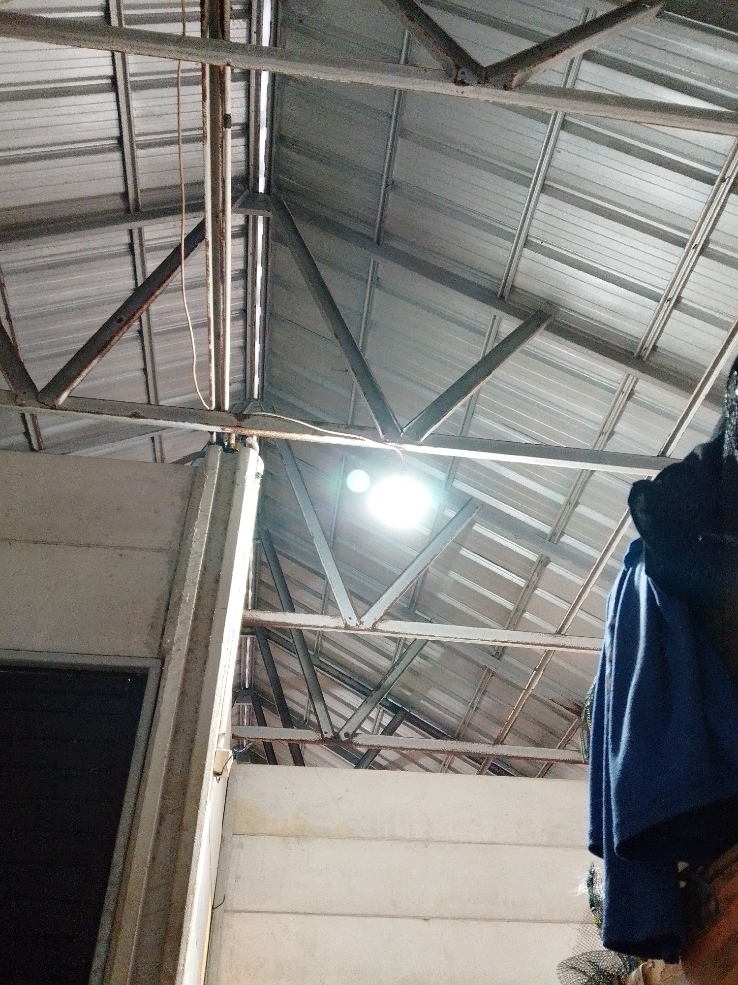A low-angle shot reveals a building interior in Bukit Ketok, Indonesia. The roof is constructed of corrugated metal supported by a framework of metal beams. A bright light fixture illuminates the scene from the ceiling. A portion of a concrete wall is visible on the left, adjacent to a dark doorway. On the right side of the image, a blue garment hangs, partially obscuring the background.