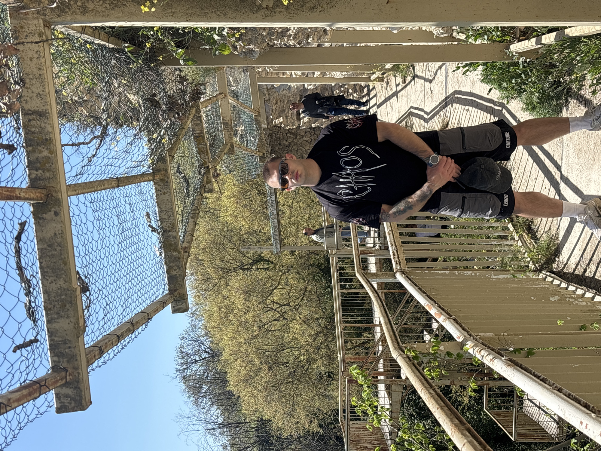 A low-angle medium shot shows a fair-skinned man in dark clothing standing on a stone stairway enclosed by rusted metal and concrete railings and a wire-mesh cage. The man is facing the viewer with dark sunglasses and has tattoos on his left arm. He is wearing a black t-shirt with white lettering, grey shorts with words printed on the side, white socks, and white shoes. He is holding a dark baseball cap in his hands, which are resting on his lap.
Behind the man, other people are walking down the stone stairway. Beyond the railings, trees with bright green leaves fill the background. The sky is blue and clear. The location appears to be an urban park.