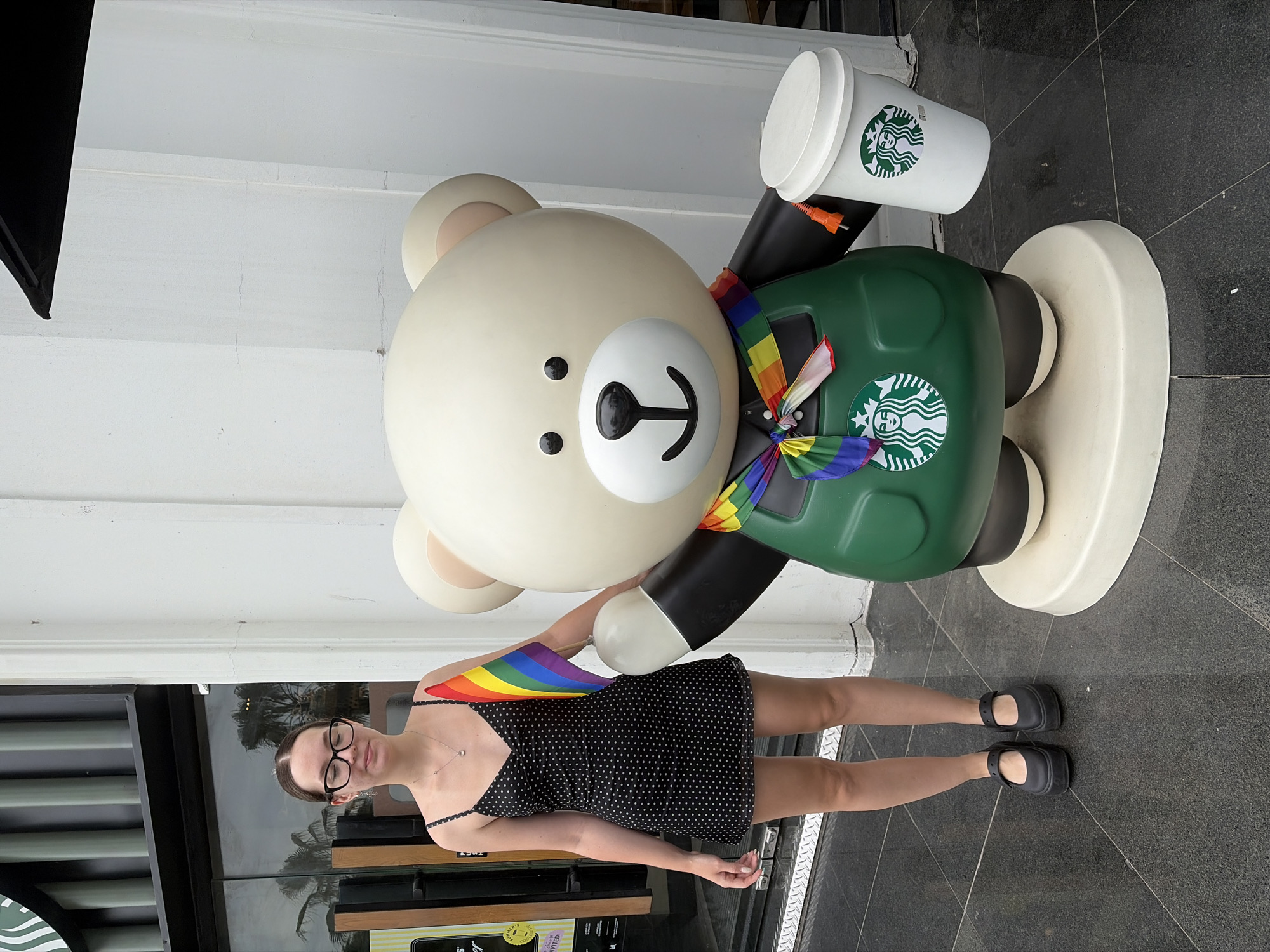 A young woman with glasses and her hair pulled back stands next to a large Starbucks bear statue. The woman is wearing a black polka dot dress and black platform shoes. She is holding a small rainbow flag in her right hand. The bear statue is wearing a green apron and a rainbow-colored scarf. It is holding a Starbucks cup in its right hand. The scene appears to be outdoors, in front of a Starbucks store, likely in Hà Nội, Vietnam, given the context. The ground is paved with dark tiles, and a white wall is visible behind the statue.