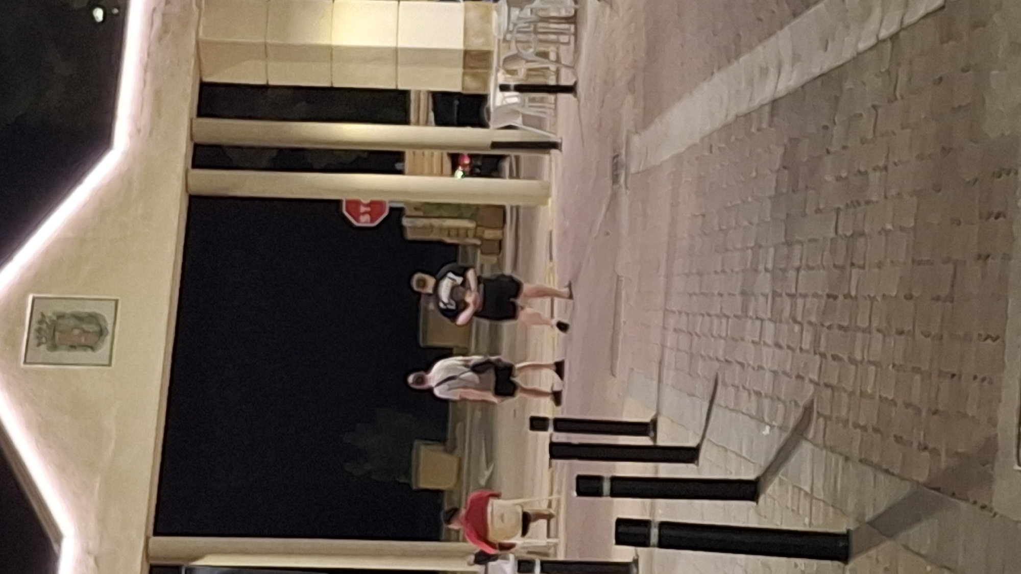 The image depicts an outdoor scene at night, likely in a public area with paving stones. In the foreground, several black bollards are visible. Further back, three men are present. One man, wearing a red shirt, is seated on a white chair to the left. Two other men are standing; one in a light-colored t-shirt and dark shorts, and another in a dark t-shirt and dark shorts. Behind the standing men, there are some potted plants and a structure with pillars, illuminated by warm lighting. A red stop sign is also visible on a pole to the right. The background is dark, suggesting a nighttime setting, and the overall environment appears to be a pedestrian area or walkway, possibly near an establishment with outdoor seating, indicated by the presence of chairs and tables. The image does not provide any specific visual cues to confirm the location in Híjar, Spain.