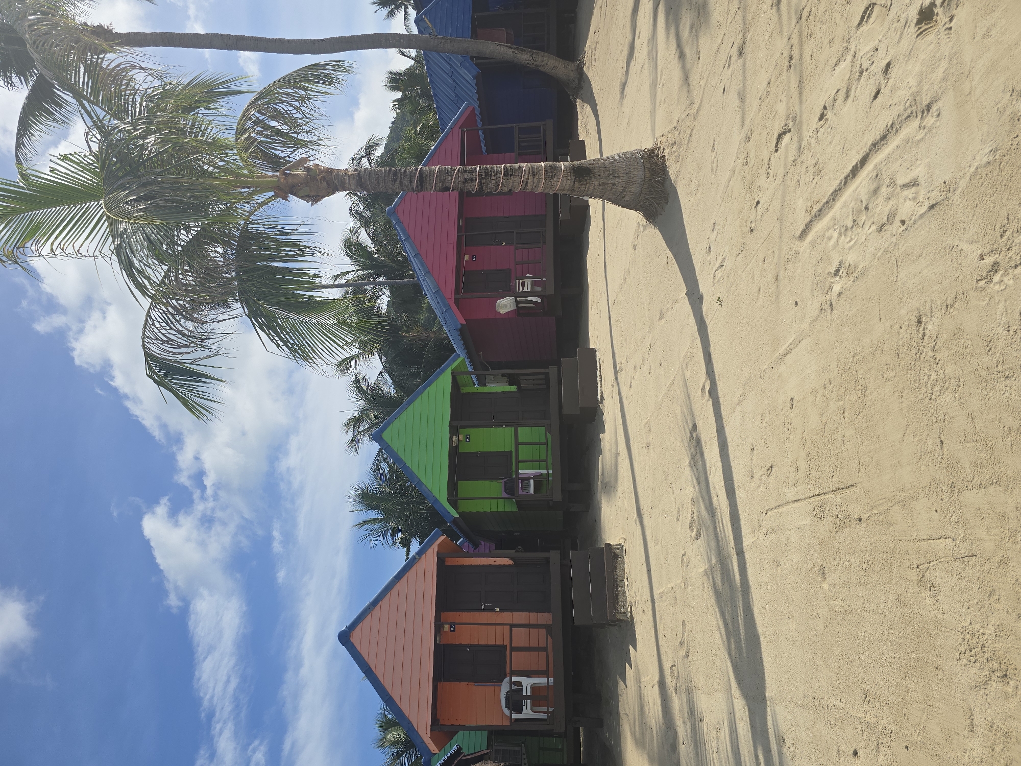 A row of brightly colored beach bungalows stands on a sandy beach under a bright blue sky with scattered white clouds. The bungalows are painted orange, green, pink, and blue, with contrasting blue roofs. Each bungalow has a small porch with a white plastic chair and a wooden door. Palm trees with lush green fronds are scattered across the scene, with the foreground dominated by footprints and shadows on the sand. The image appears to be taken in Baan Lamai, Thailand, a location known for its beaches and tourist accommodations.