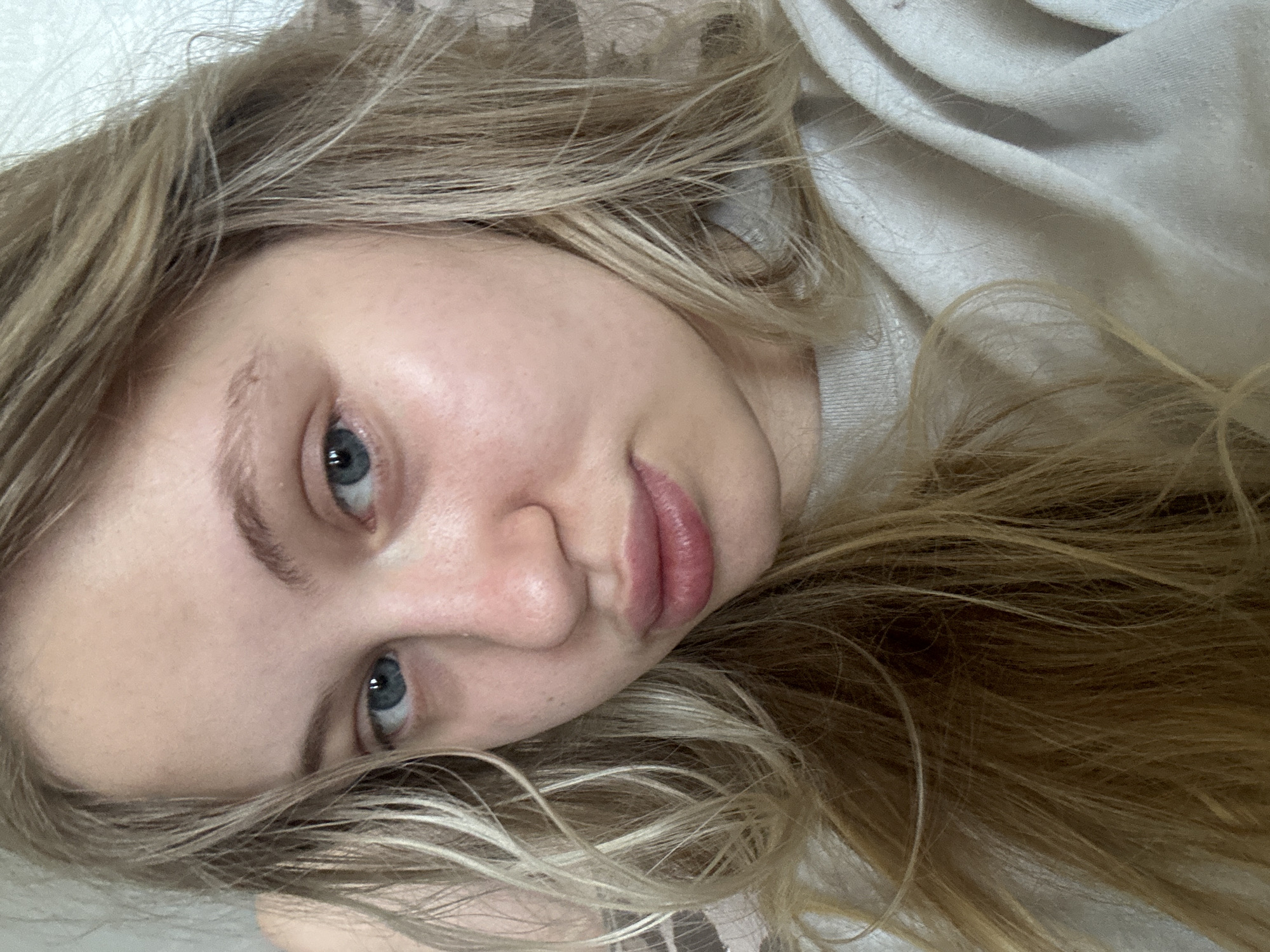 The image is a close-up, slightly angled portrait of a young Caucasian woman with blonde hair and blue eyes, looking directly at the camera. She is wearing a light-colored, possibly grey or beige, crew-neck sweatshirt. The background is indistinct and appears to be an indoor setting. There are no identifiable landmarks or elements that would indicate a specific location within the city of Miass, Russia. The lighting suggests an indoor environment, possibly natural light coming from a window. The woman's expression is neutral.