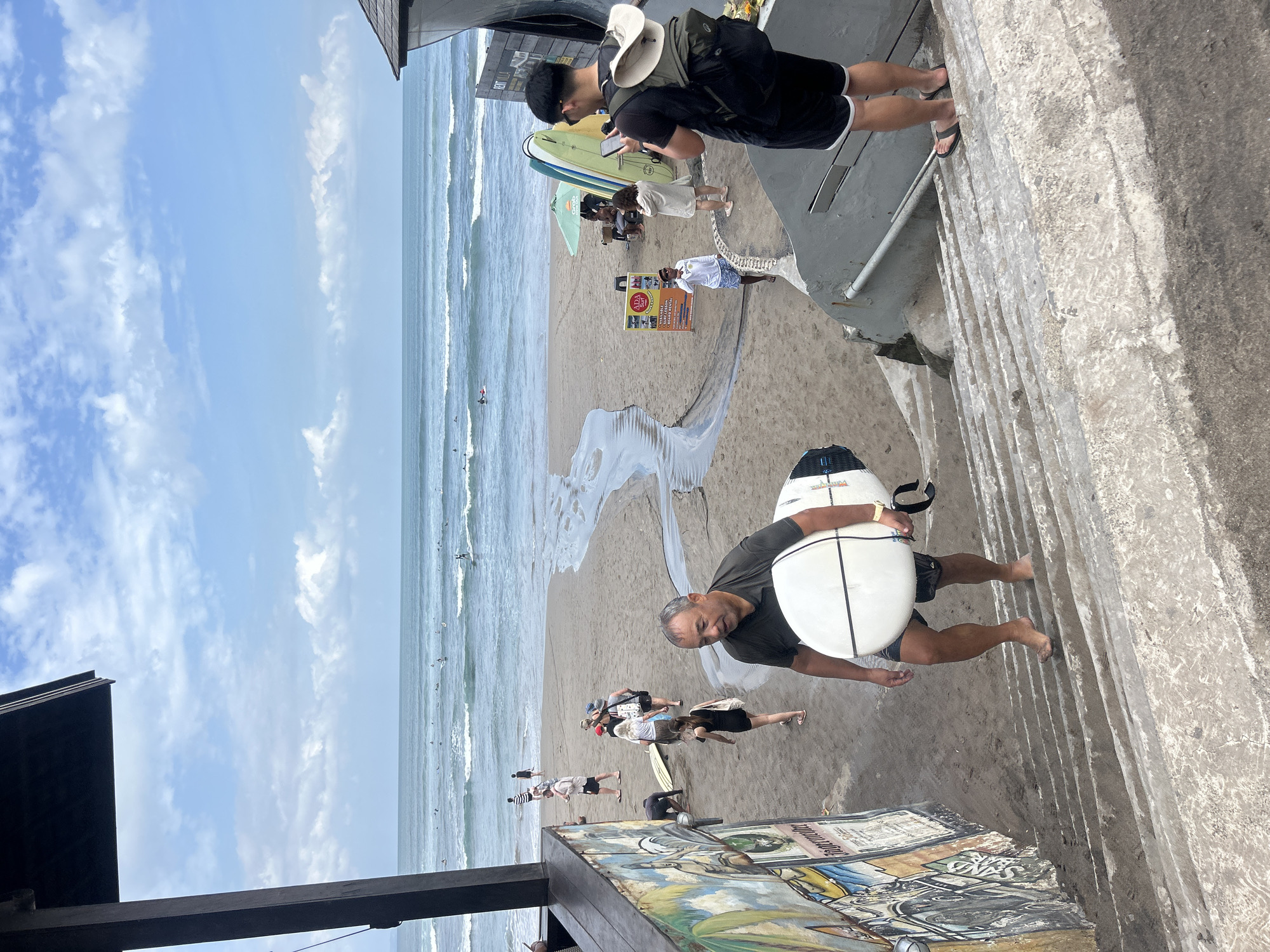 This image depicts a scene on a beach in North Kuta, Indonesia, with a focus on a man carrying a surfboard up a set of concrete stairs. The man, who appears to be of Asian descent and middle-aged, is wearing a dark t-shirt and dark shorts. He is barefoot and holding a white surfboard under his left arm. The stairs are wide and appear to be made of concrete, with some visible dirt and wear.
In the background, the beach is sandy, and the ocean stretches out to the horizon under a partly cloudy sky. There are several other people on the beach and in the water, many of whom are surfers. A small stream of water is visible flowing across the sand towards the ocean. To the left of the stairs, there is a wall with colorful artwork. To the right, a darker structure, possibly a building or a shaded area, is visible. Another person, a young man wearing a black t-shirt, shorts, and a backpack, is standing on the stairs above the man with the surfboard, looking at his phone. Other individuals, including women and children, are scattered across the beach and near the water. Some surfboards are propped up on the sand. A sign with text, possibly advertising rentals, is also present. The overall impression is a busy beach environment with recreational activities taking place.