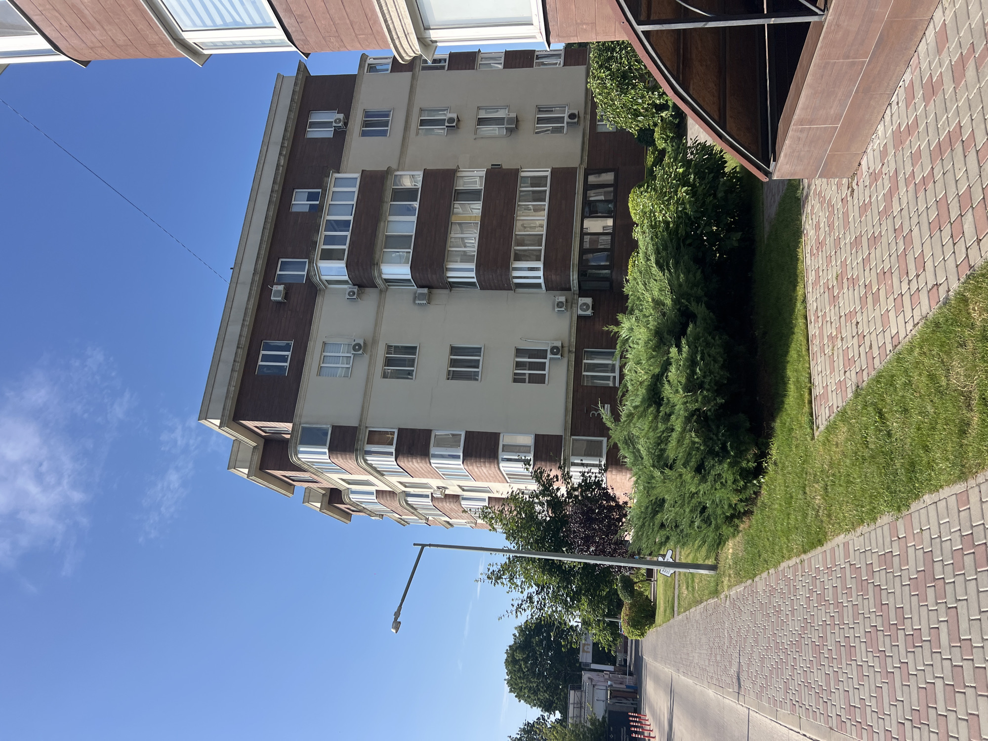 The image depicts a sunny day in Kryzhanivka, Ukraine, with a clear blue sky. A multi-story apartment building with light beige and dark brown facade elements and numerous windows and air conditioning units is prominently featured. To the left of the building, a paved sidewalk with a brick pattern runs parallel to a road. A street lamp stands on the sidewalk. Green grass and lush bushes, including a large juniper, are visible in front of the building and along the sidewalk. The building on the right, partially visible, has a curved facade and a similar color scheme. A covered structure with a wooden frame and a dark roof is situated in the foreground on the right. The scene appears to be a residential area.
