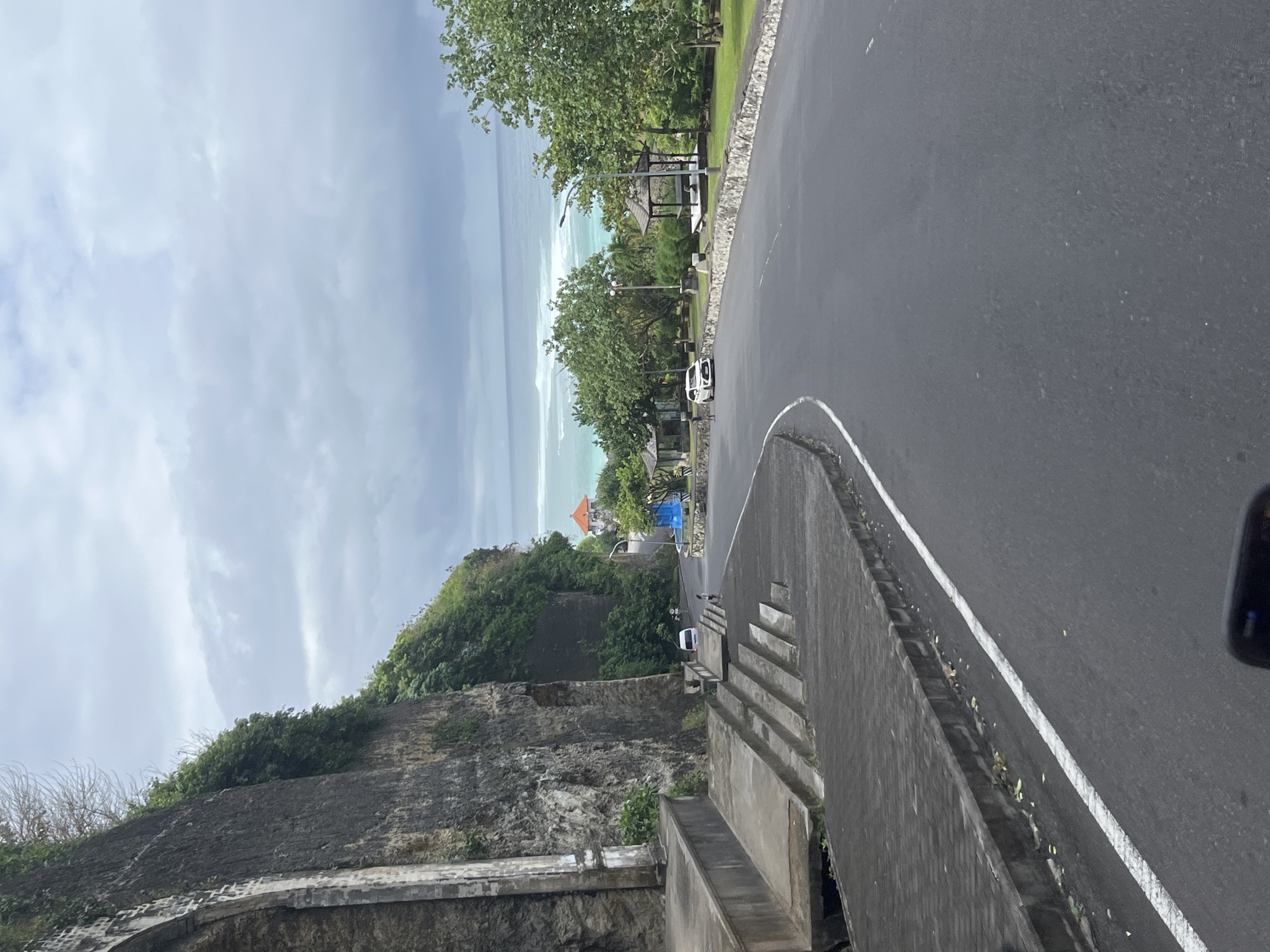 The image depicts a view from a high vantage point in Kuta Selatan, Indonesia. The location appears to be near Uluwatu Temple, given the rocky cliffside and ocean view. The scene shows a paved road curving along the edge of a cliff. On the left, there is a steep, rocky cliff with vegetation clinging to its surface. Below the road, there is a building with a red roof and blue construction material. The ocean extends into the distance with white waves breaking along the shoreline. The sky is overcast with gray clouds. On the right side of the road, there are trees and shelters. A few cars are visible along the road. The photo is taken at an angle. A concrete staircase and wall are in the foreground of the photo.