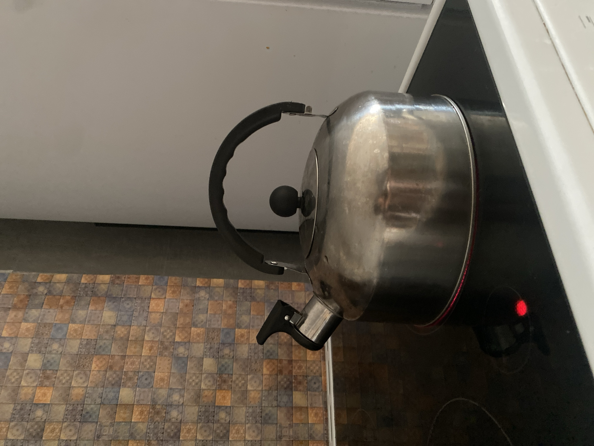 An overhead shot of a silver metal stovetop kettle with a black handle and lid is displayed on a black electric stovetop. The kettle is centered in the frame, and its rounded body reflects the ambient light, showing some minor wear and tear. A red indicator light is visible on the stovetop beneath the kettle, suggesting it is currently heating. In the background, a tiled wall with a mosaic pattern of various shades of brown, blue, and orange is partially visible on the left, while a white refrigerator or wall panel occupies the right side of the frame. The scene appears to be set in a kitchen. This image was taken in Novosibirsk, Russia.