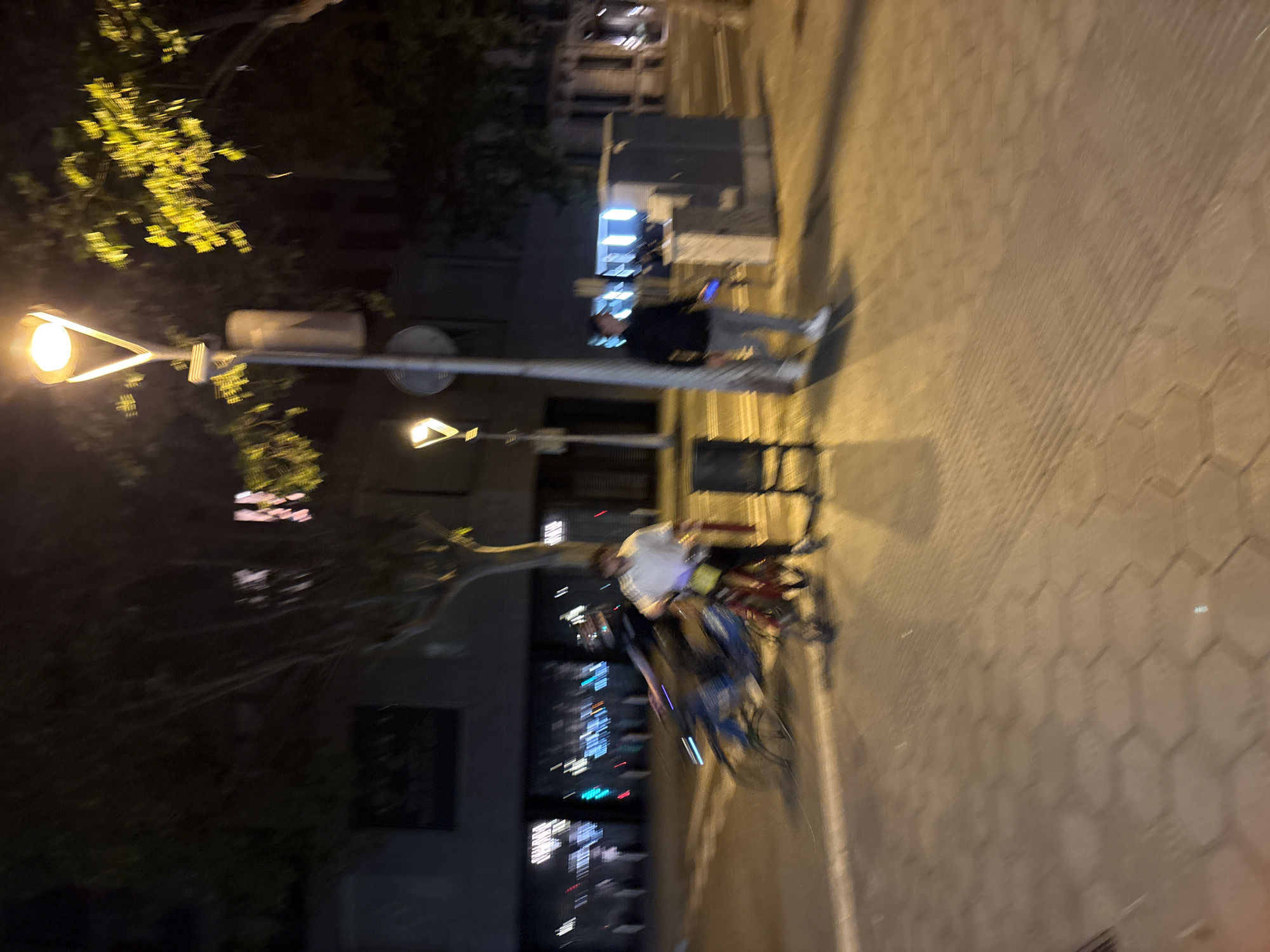 The image captures a night scene in Barcelona, Spain. A street lamp illuminates the surroundings, casting light on a person seated on a bicycle. Buildings and street lights blur in the background, typical of an urban environment. On a pedestrian bridge, an individual is seen standing and using a cell phone. The ground is covered with geometric cobblestones.
