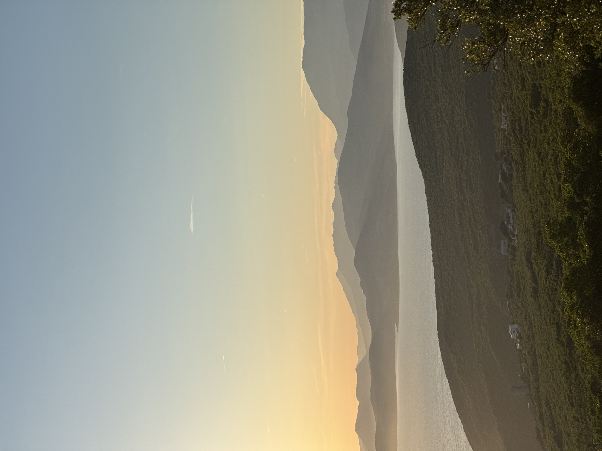 This is a vertical, wide-angle, eye-level, full shot of a serene coastal landscape at dusk in Kovačko Polje, Montenegro. The scene is dominated by layers of silhouetted mountains receding into the distance, their peaks softened by the fading light. A calm body of water, likely the Adriatic Sea, occupies the middle ground, reflecting the soft hues of the sky. The foreground is a lush, green hillside covered in dense vegetation, with a few scattered white buildings nestled amongst the trees, suggesting a small settlement. The sky transitions from a pale, soft yellow near the horizon, where the sun has recently set, to a clear, light blue overhead. A faint wisp of a cloud and two faint contrails from aircraft are visible in the upper sky. The overall mood is peaceful and tranquil, characterized by muted colors and soft light.