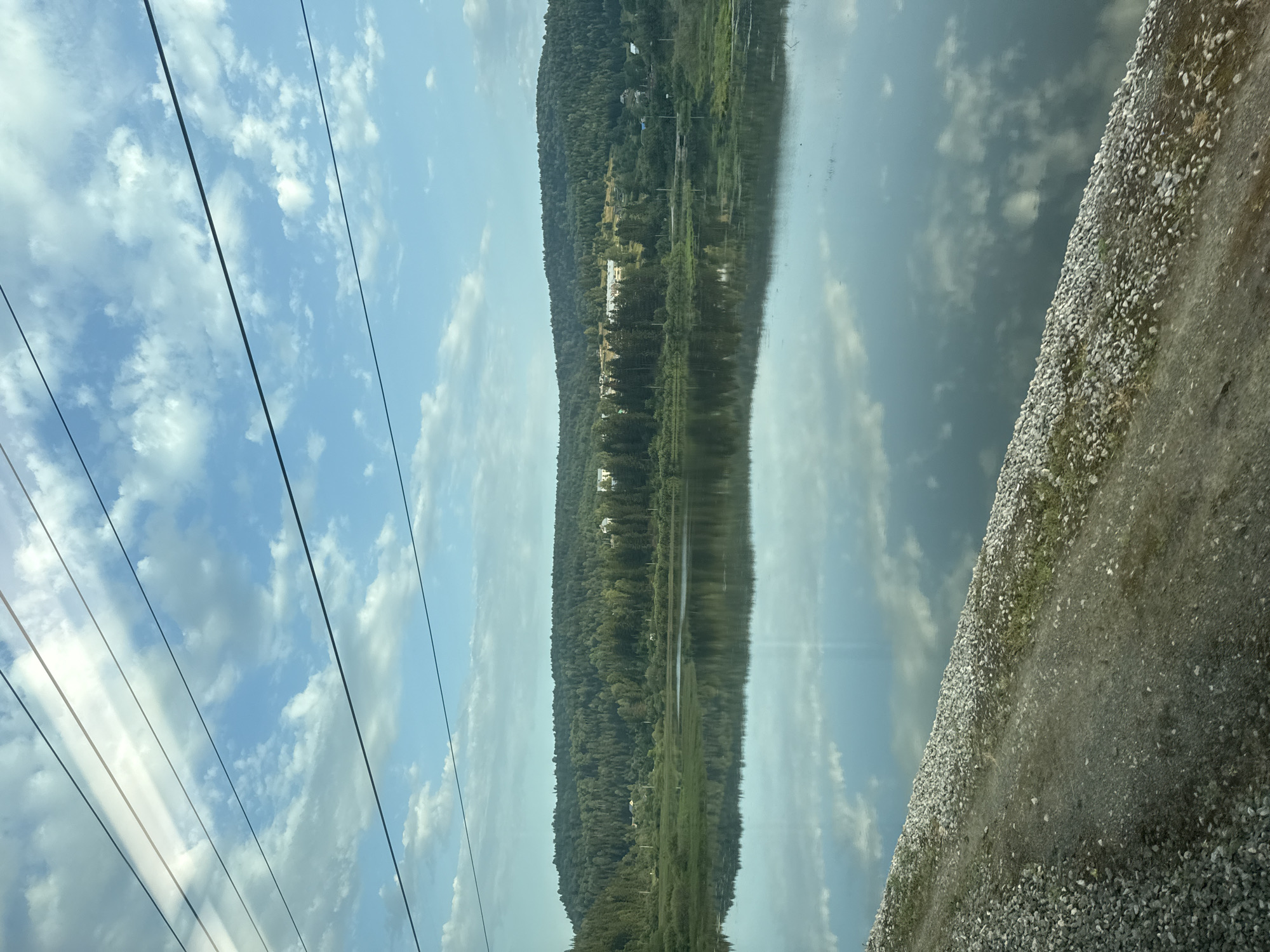 A body of water reflects a treed hillside under a cloudy sky. The water is calm and mirrors the sky and the landscape. The hillside is covered in dense evergreen trees. There are some buildings visible on the hillside. The foreground shows a gravelly bank leading down to the water's edge. Overhead, power lines cross the sky. This scene is located on the outskirts of Minyar, Russia, near the Ural River.
