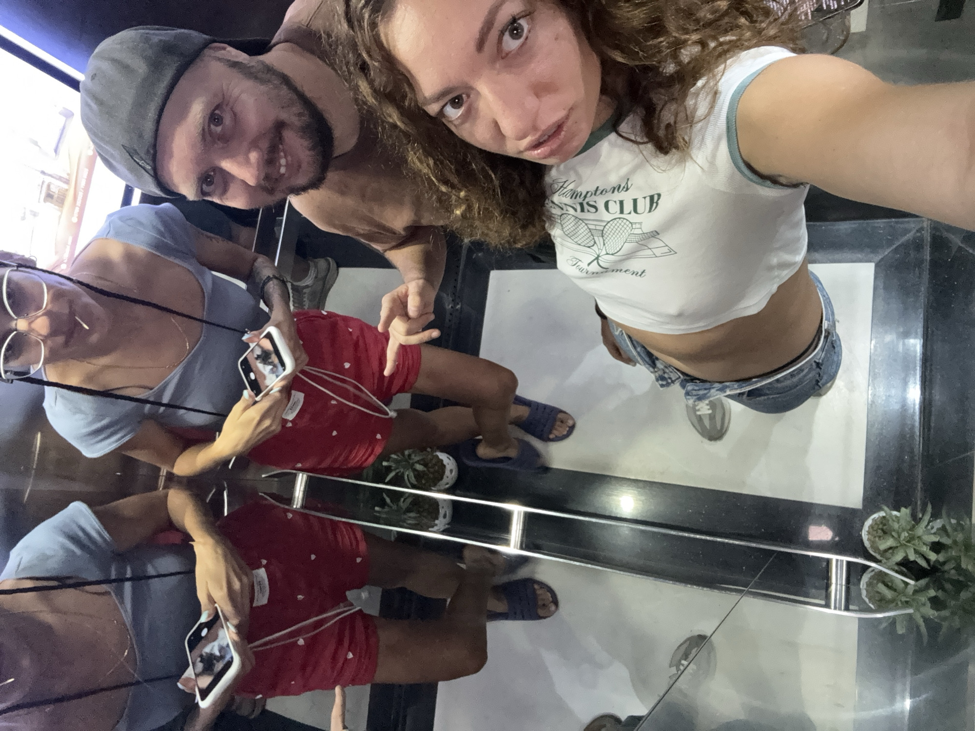 In an elevator, two women and a man are taking a selfie. The woman in the foreground is looking at the camera, her curly brown hair framing her face. She is wearing a white crop top with the words "Hampton's Tennis Club" and "Tournament" printed on it, and denim shorts. The man next to her, wearing a brown t-shirt and a grey baseball cap, is also looking at the camera with a smile. Behind them, another woman, wearing a blue t-shirt and red shorts with white hearts, is holding a smartphone and looking at it. The elevator has reflective walls, showing the reflections of the people and the plants in pots. The floor is white with black lines and a metal railing. The scene is indoors and appears to be in a modern building, possibly a hotel or apartment complex. There is no specific indication of the city of Hanoi, Vietnam.