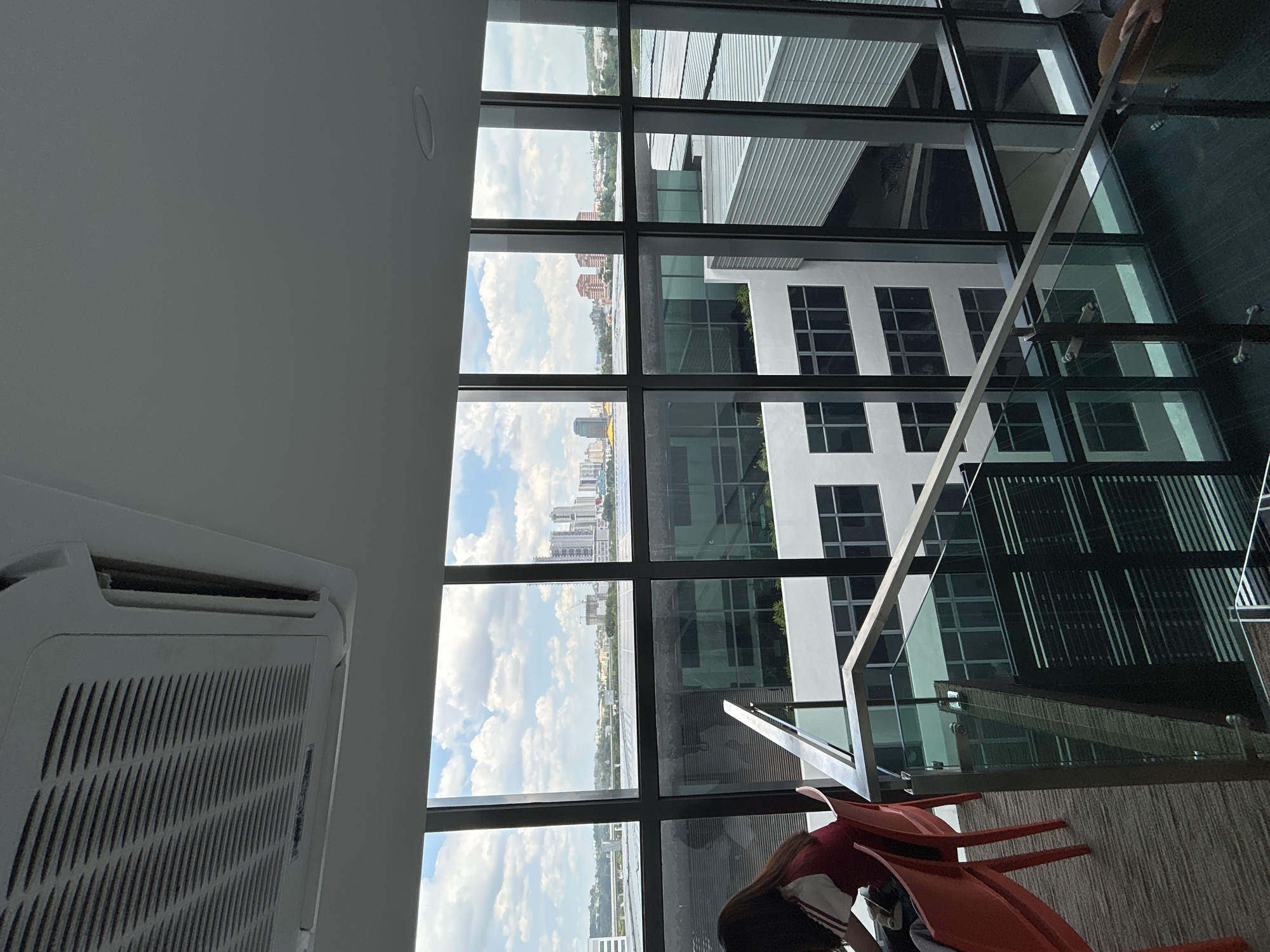 The view from a high-rise building in Kuala Lumpur, Malaysia, is captured through a large window with a black grid frame. The cityscape includes other buildings, some modern and some older, with a cloudy sky overhead. Inside the building, an air conditioning unit is visible near the ceiling. In the lower part of the frame, a person with long dark hair is partially visible, seated on a red chair. The interior includes a glass railing and part of a staircase.