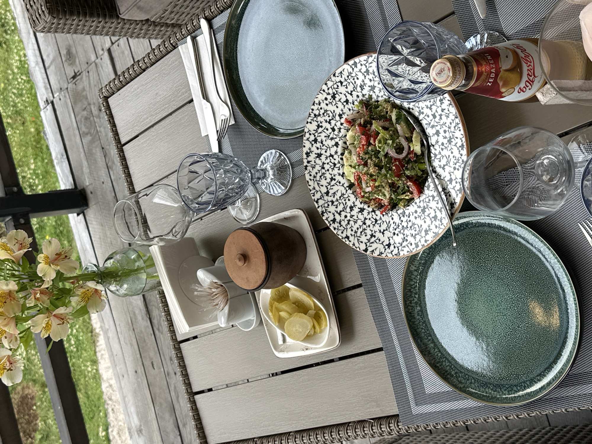The image captures a table setting, likely outdoors, with a focus on a meal setup. The table is made of a grey, textured material, possibly wood or a composite. The setting includes plates, cutlery, glasses, and a bottle of juice, all arranged on a striped placemat. There is a plate of salad with visible ingredients like tomatoes, greens, and onions. Another plate holds sliced lemons or pickled vegetables, alongside a small, brown, lidded container. There is a vase of light-colored flowers towards the edge of the table. The scene is set against a backdrop of green grass and a wooden structure, suggesting a patio or garden setting. No people are visible in the frame. The city of Tibeta, Georgia is not discernible from the visual information provided.