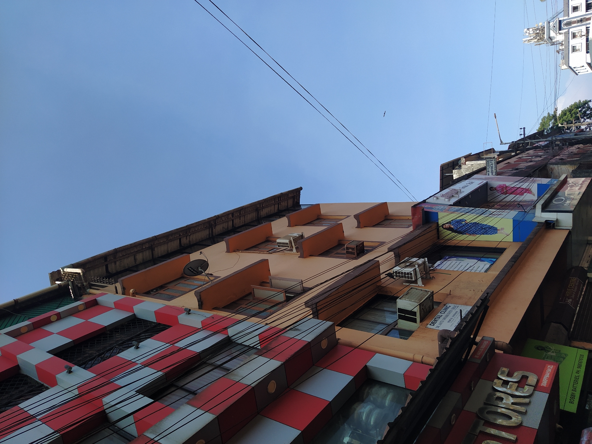 The image captures a low-angle perspective of a street scene in Mapusa, India, dominated by a view of multi-story buildings. The buildings exhibit varying architectural styles, from a red and grey checkered facade to a peach-colored building with air conditioning units and satellite dishes. Electrical wires crisscross the sky. Signage is visible on the buildings, including "DENTAL CLINIC," "STORES," and "ADVOCATE." Bobby Batteries are also advertised. A clear blue sky fills the upper portion of the frame, in which a small bird is seen. The buildings are stacked closely together, characteristic of urban density.