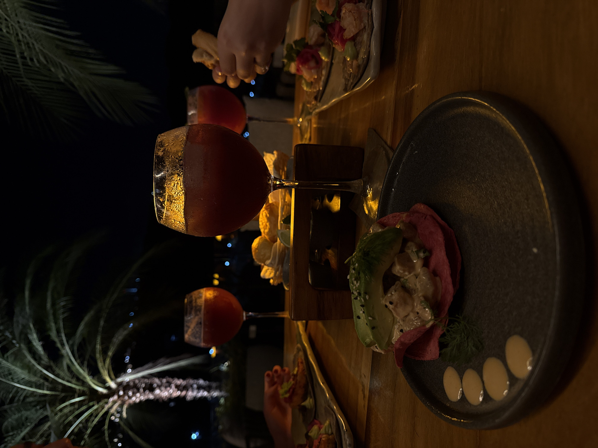 The image shows a dimly lit outdoor setting, likely in the evening, given the dark background. A wooden table is the central focus, displaying a variety of dishes and drinks. Two glasses filled with a red liquid are prominent, positioned amidst the food items. The food consists of what appears to be a taco with avocado and some white sauce dots on the side, alongside other dishes of an indiscernible nature. Two palms tree are clearly seen in the background. The scene suggests a meal at a restaurant in Puerto Marqués, Mexico, characterized by its outdoor setting and a variety of dishes. The overall atmosphere is relaxed, indicating an enjoyable dining experience.