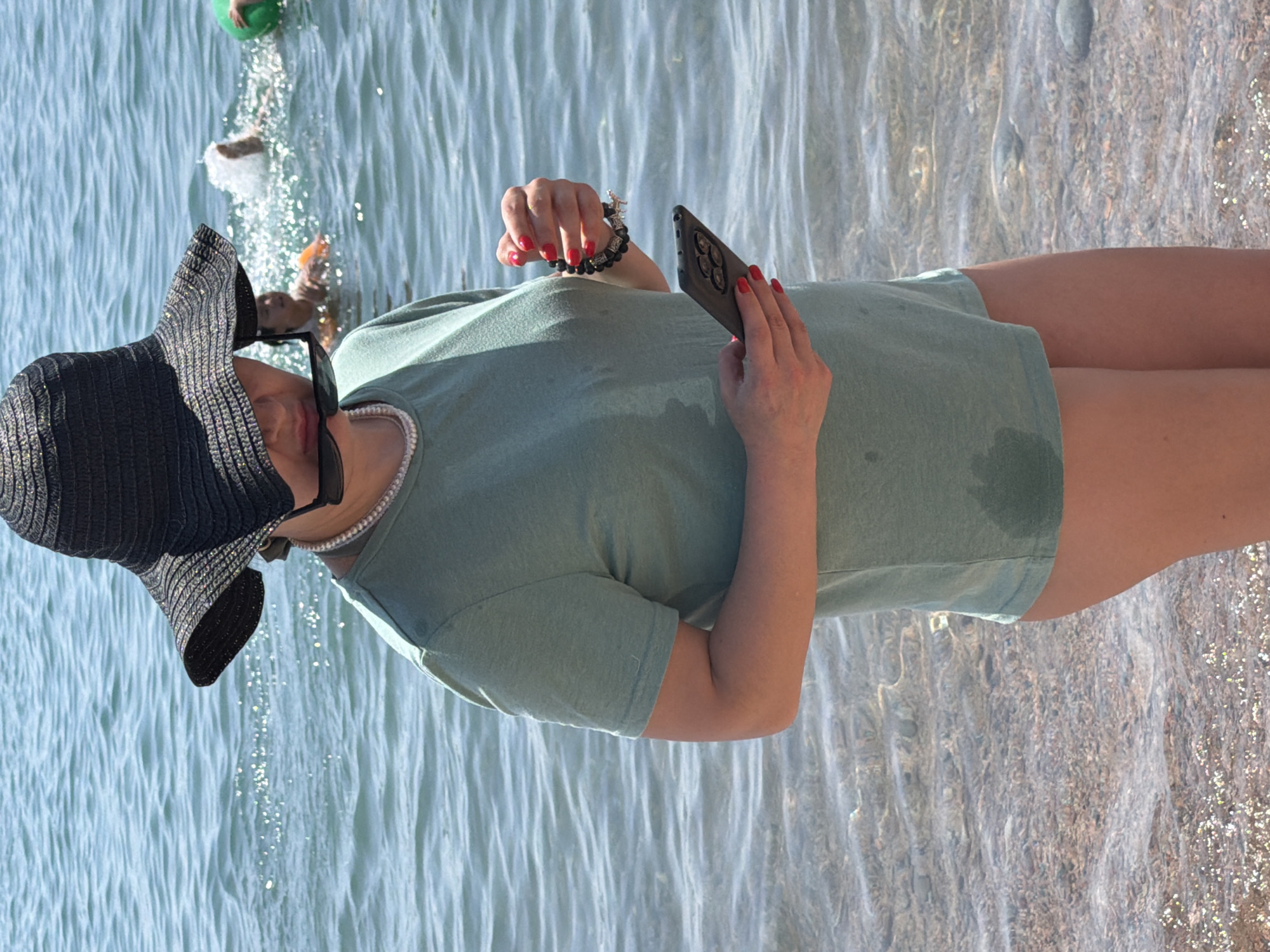 A woman wearing a black wide-brimmed hat and a pale green t-shirt stands at the edge of the water. She holds a black smartphone in her right hand and a bracelet made of dark beads and silver charms in her left. Her fingernails are painted red. In the background, two children are swimming in the blue water, one of them holding a green inflatable ring. The woman's t-shirt is damp in the front.
