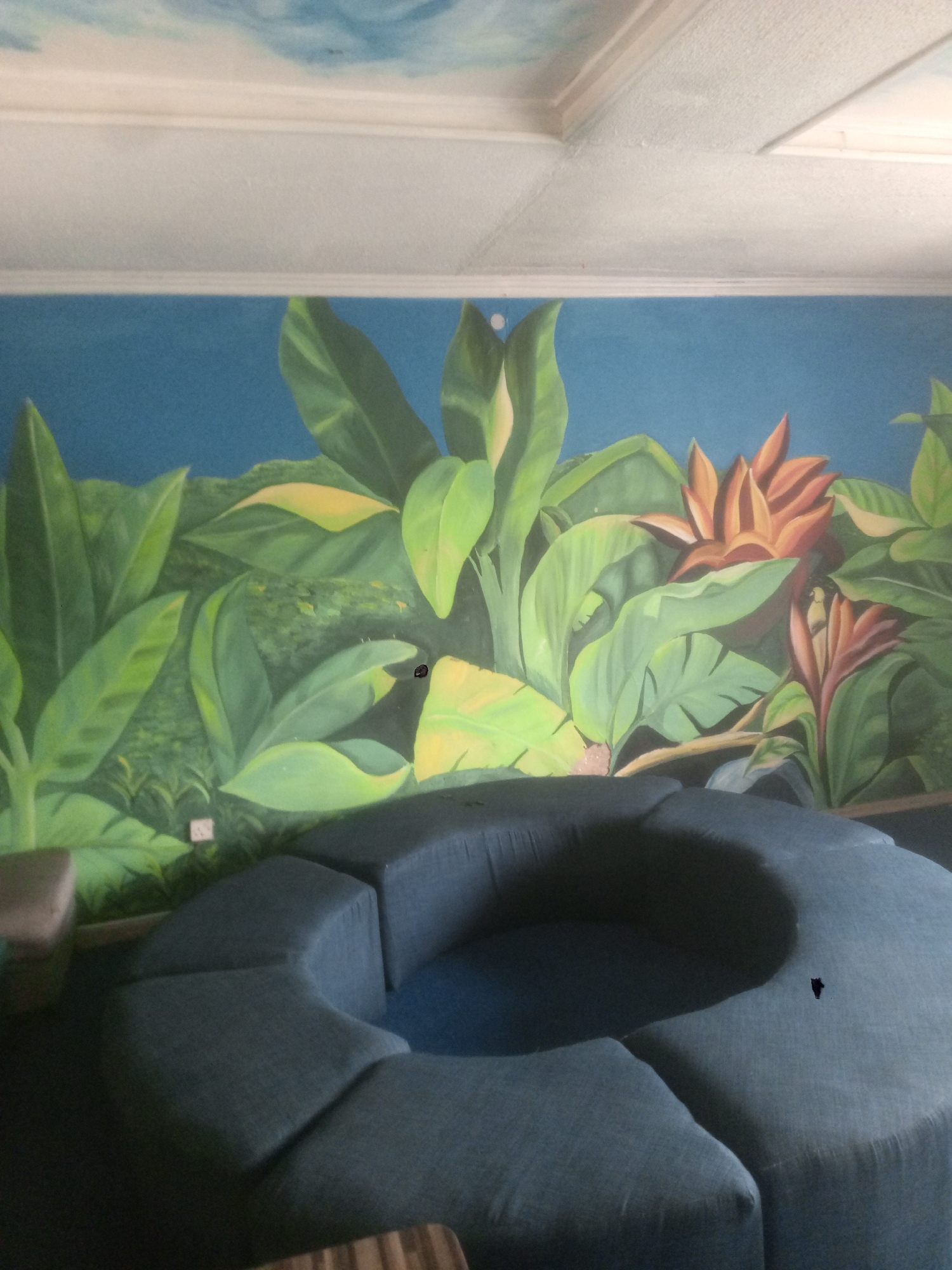 The image depicts a room featuring a semi-circular blue couch in the foreground, set against a wall mural displaying tropical foliage and flowers. The environment suggests an indoor space, likely a lounge or waiting area. The ceiling is white, with a painted blue sky pattern visible in one corner. The location is Abuja, Nigeria.