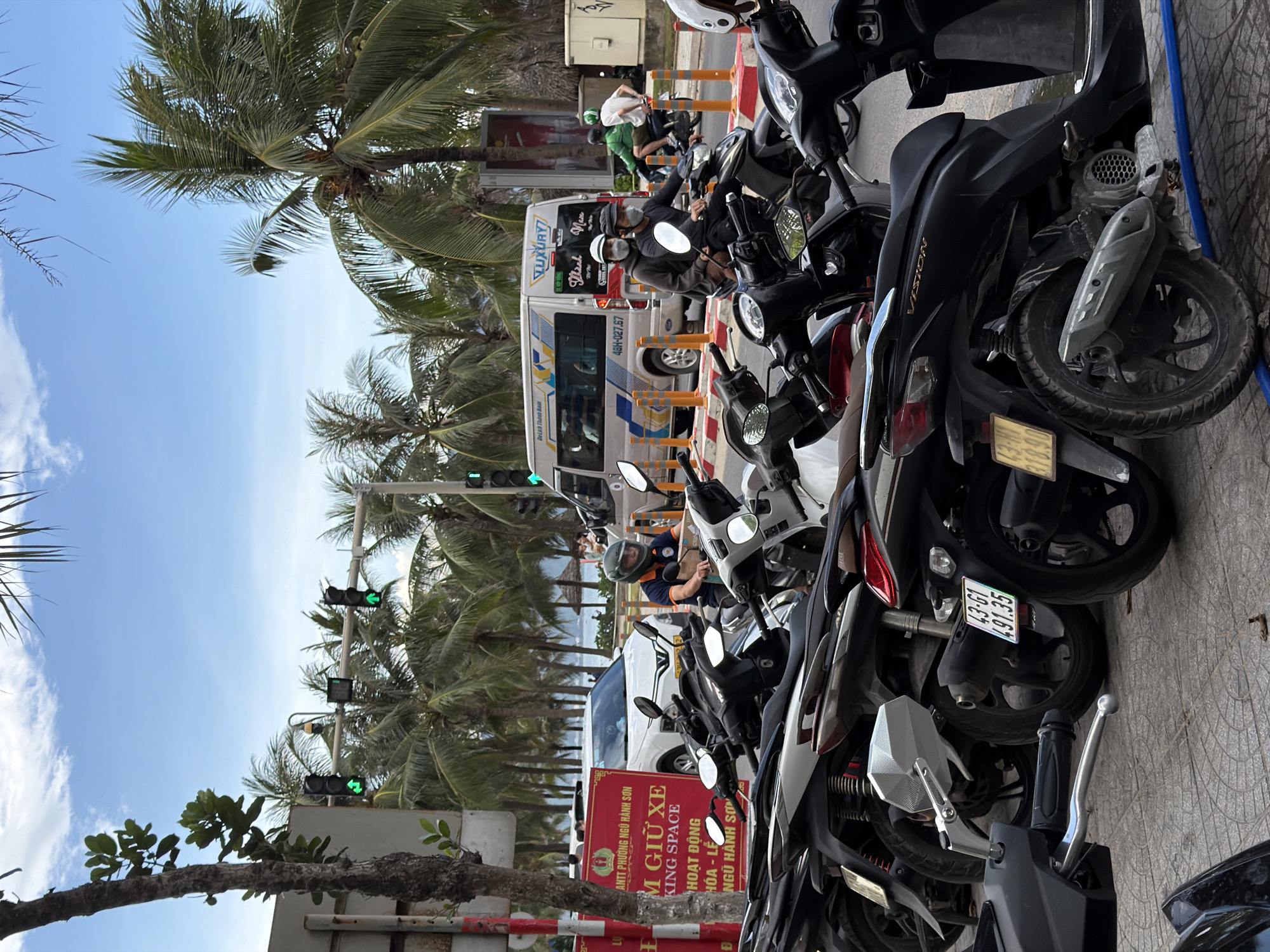 This is a street scene in Đà Nẵng, Vietnam, likely in the daytime given the bright sky and shadows. The foreground is dominated by a row of parked motorcycles, with several people on them, some wearing helmets. A white van with "LUXURY" and "Thanh Nam" visible on its side is parked behind the motorcycles, with people visible inside. The background features a line of palm trees, suggesting a coastal or tropical environment. Traffic lights are visible, with green signals illuminated. A red sign with Vietnamese text, including "M GIỮ XE" (which translates to "Parking") and "KING SPACE", is also visible, indicating a parking area. The overall impression is a busy street scene with many vehicles and people. The weather appears to be clear and sunny.