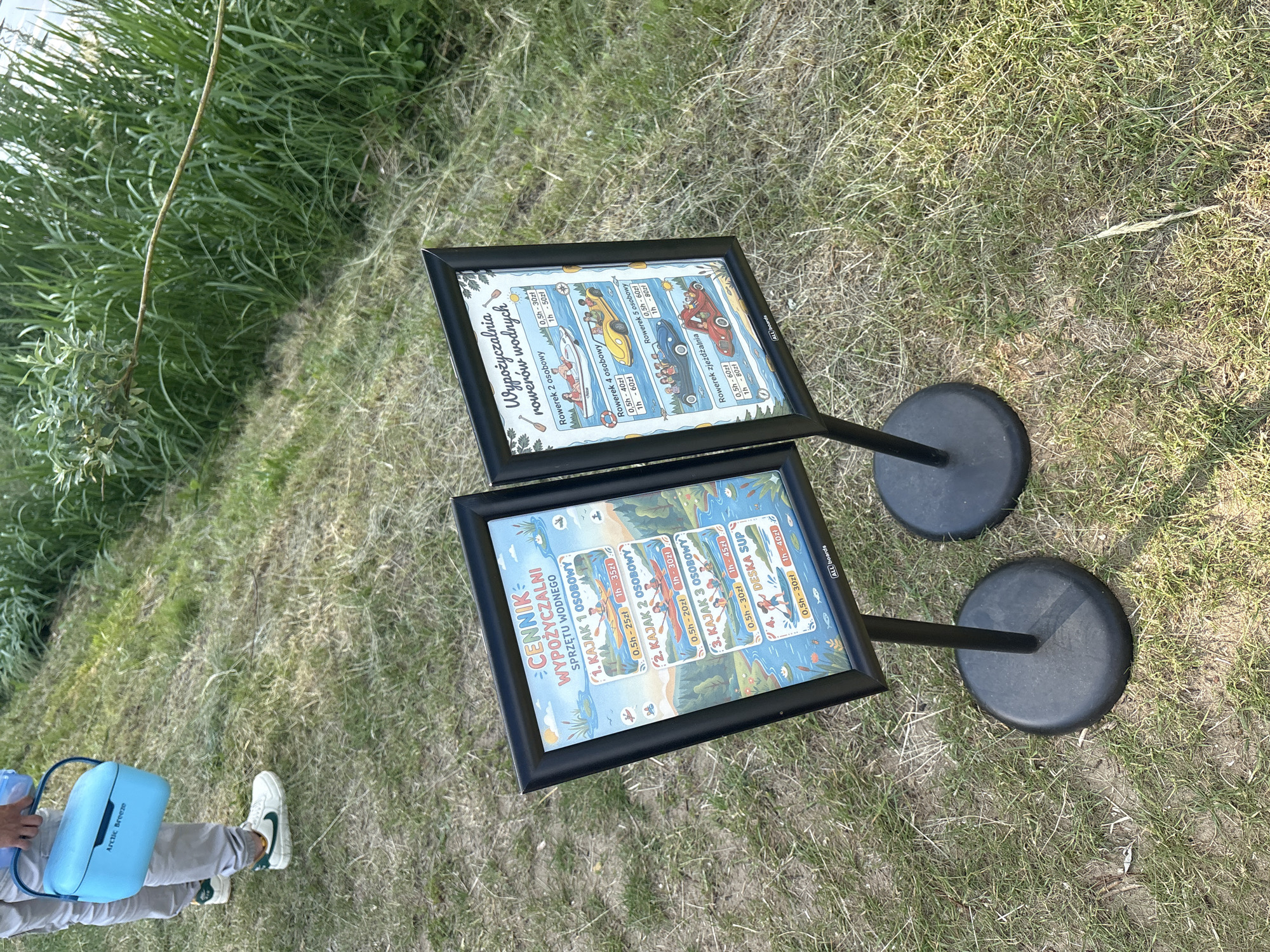 The image shows two framed signs on stands outdoors, likely in the city of Kobyla Góra, Poland, near a body of water, as indicated by the context of the signs which detail rental prices for water sports equipment. The sign on the left is titled "CENNIK WYPOŻYCZALNI SPRZĘTU WODNEGO" (Price list for water equipment rental) and lists prices for kayaks and a SUP board. The sign on the right is titled "Wypożyczalnia rowerów wodnych" (Water bike rental) and displays prices for different types of water bikes. In the background, there is grassy terrain and lush green vegetation. In the foreground, a person is partially visible on the left, holding a light blue cooler bag labeled "Arctic Breeze." The overall scene suggests a rental service for recreational water activities.