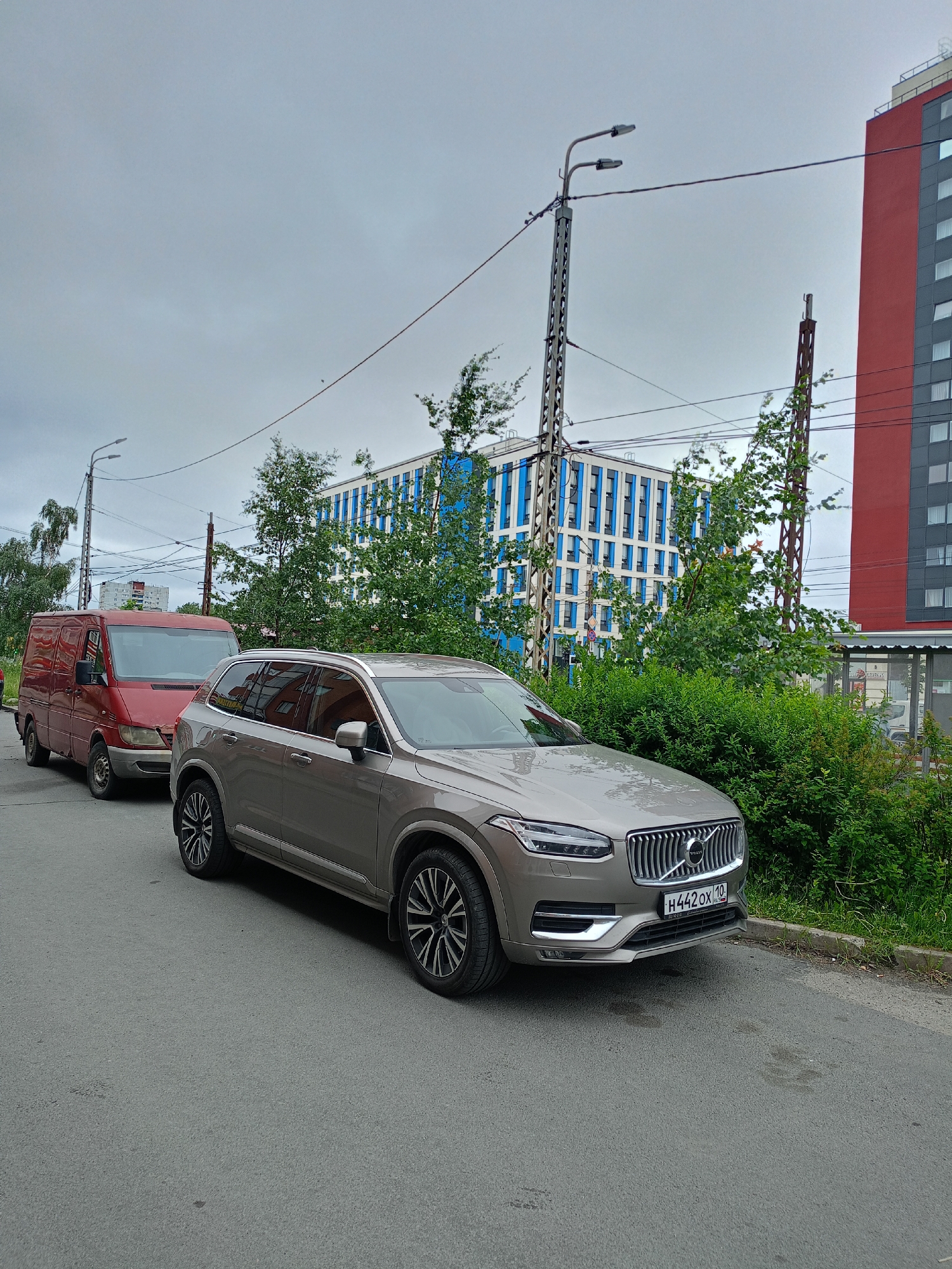 An exterior, eye-level, medium shot captures two cars parked on a paved road in front of buildings under a cloudy sky. On the left, a red panel van is parked parallel to the curb. To its right and slightly closer to the viewer, a silver Volvo SUV is also parked parallel to the curb, facing right. The SUV's front license plate, bearing the characters "H442OX 190", is visible. Behind the cars, a modern building with blue and white vertical accents stands to the left, while a tall, red-colored building with dark gray windows is to the right. Lush green trees and bushes are situated between the road and the buildings. Power lines crisscross the scene, supported by metal poles, one of which has two streetlights attached. The ground is a gray asphalt surface with a visible curb and some scattered debris.