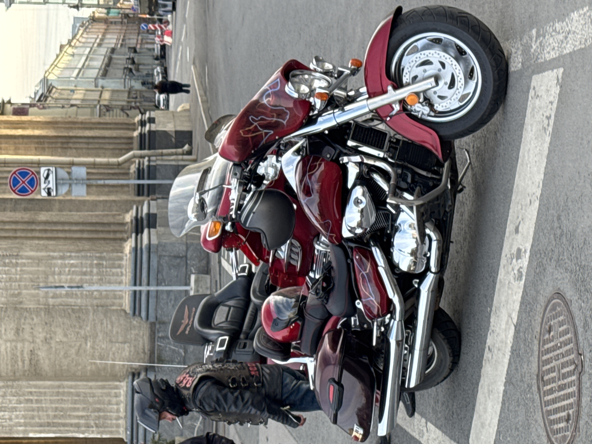 The image shows two maroon motorcycles parked on the side of a street, with a man standing next to them. The motorcycles are parked in a parking space marked by a white line on the pavement. The background shows a building with columns, and a street sign with a blue and red 'no parking' symbol. The scene is likely in Saint Petersburg, Russia, based on the architectural style of the building in the background, and the style of the street sign. The image shows motorcycles parked next to the building.