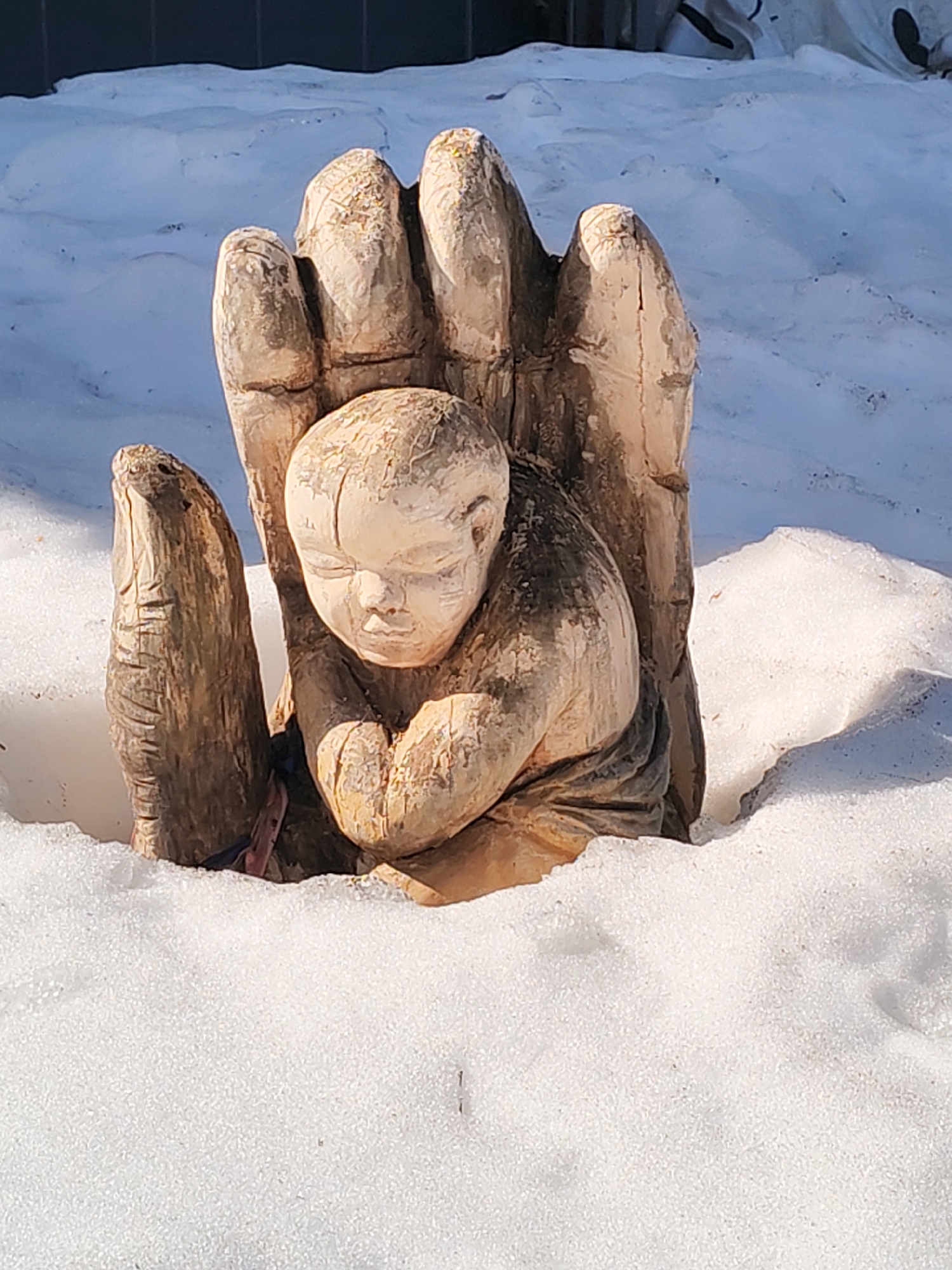 A wood carving depicting a baby resting in a hand is partially buried in snow. The location is Chaikovsky, Russia.