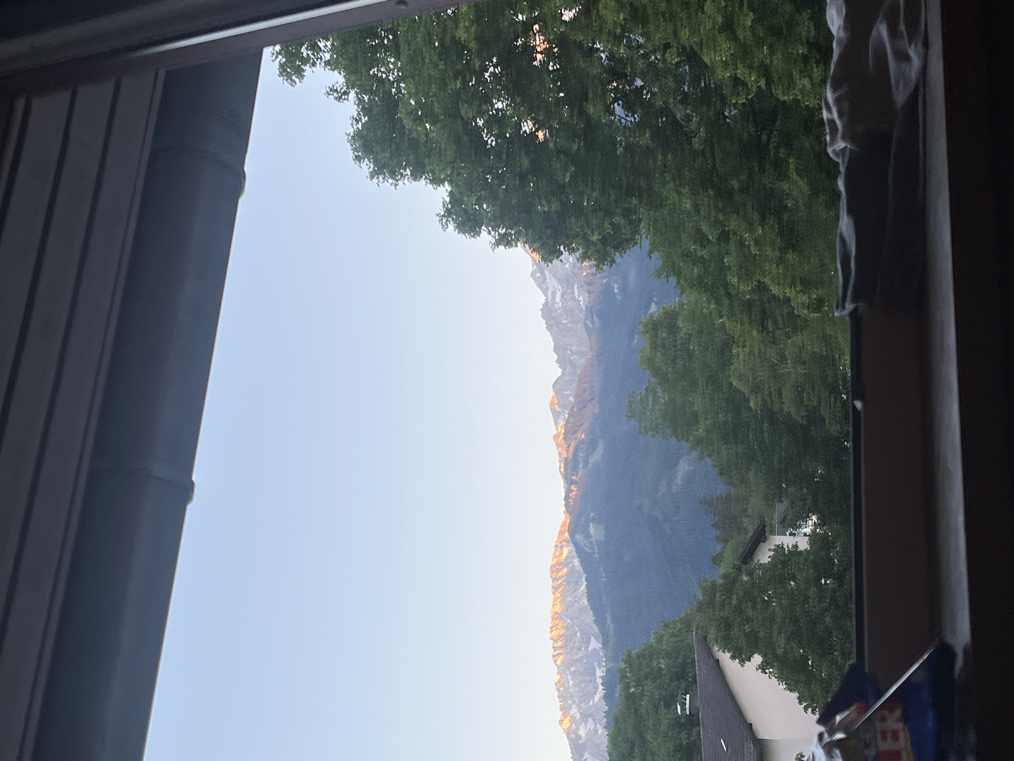 A view from a window in Garmisch-Partenkirchen, Germany, reveals a scenic landscape. The frame of the window is visible, with a metal exterior on the left and a darker wooden frame on the right. Outside the window, lush green trees partially obscure the view of the mountains in the distance. The mountain range features rocky peaks, some covered with snow, illuminated by a warm, golden light, likely from the rising or setting sun. A building with a white facade is visible among the trees. The sky is a clear, pale blue. The image is rotated, with the horizontal scene presented vertically.