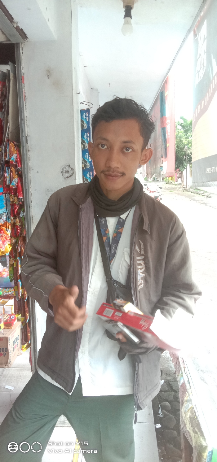 A young adult male, appearing to be of Indonesian ethnicity, stands under a covered walkway of a building, likely a small shop or "warung," in Sebaung, Indonesia. He has short, dark, wavy hair, dark eyes, and a thin mustache. He wears a dark brown zip-up jacket over a white long-sleeved shirt, dark green pants, and a black neck gaiter. A black shoulder bag with a colorful "SUKUN" branded lanyard is visible. In his right hand (viewer's left), he holds several packs of cigarettes, including red and yellow-boxed varieties, along with a silver-colored object, possibly a lighter. His left hand (viewer's right) is partially raised with an open palm. To his left, shelves stocked with various packaged snack foods and brightly colored items are visible within a storefront. Above him, a single bare light bulb is suspended from a white ceiling. To his right and behind him, a street scene is visible, featuring loose rocks in a gutter, green foliage, and a partial view of a red motorcycle, all under daylight conditions.