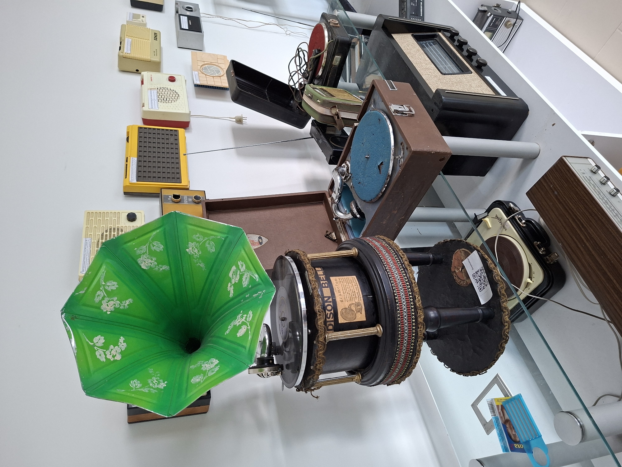 The image depicts a collection of vintage audio equipment, including gramophones and radios, displayed on white shelves against a white wall. Prominently featured is a large, green, flared gramophone horn with floral detailing, connected to a black cylindrical base with decorative trim. To the right, several portable radios of various colors and designs are mounted on the wall. On a glass shelf below, a brown suitcase-style record player with a blue turntable is open, next to other portable record players and a larger radio with a fabric speaker grille. A smaller, wooden-cased radio is visible on a lower shelf. The scene suggests a museum exhibit or a private collection of historical electronics. The location is identified as Ialoveni, Moldova. No people are present in the image.