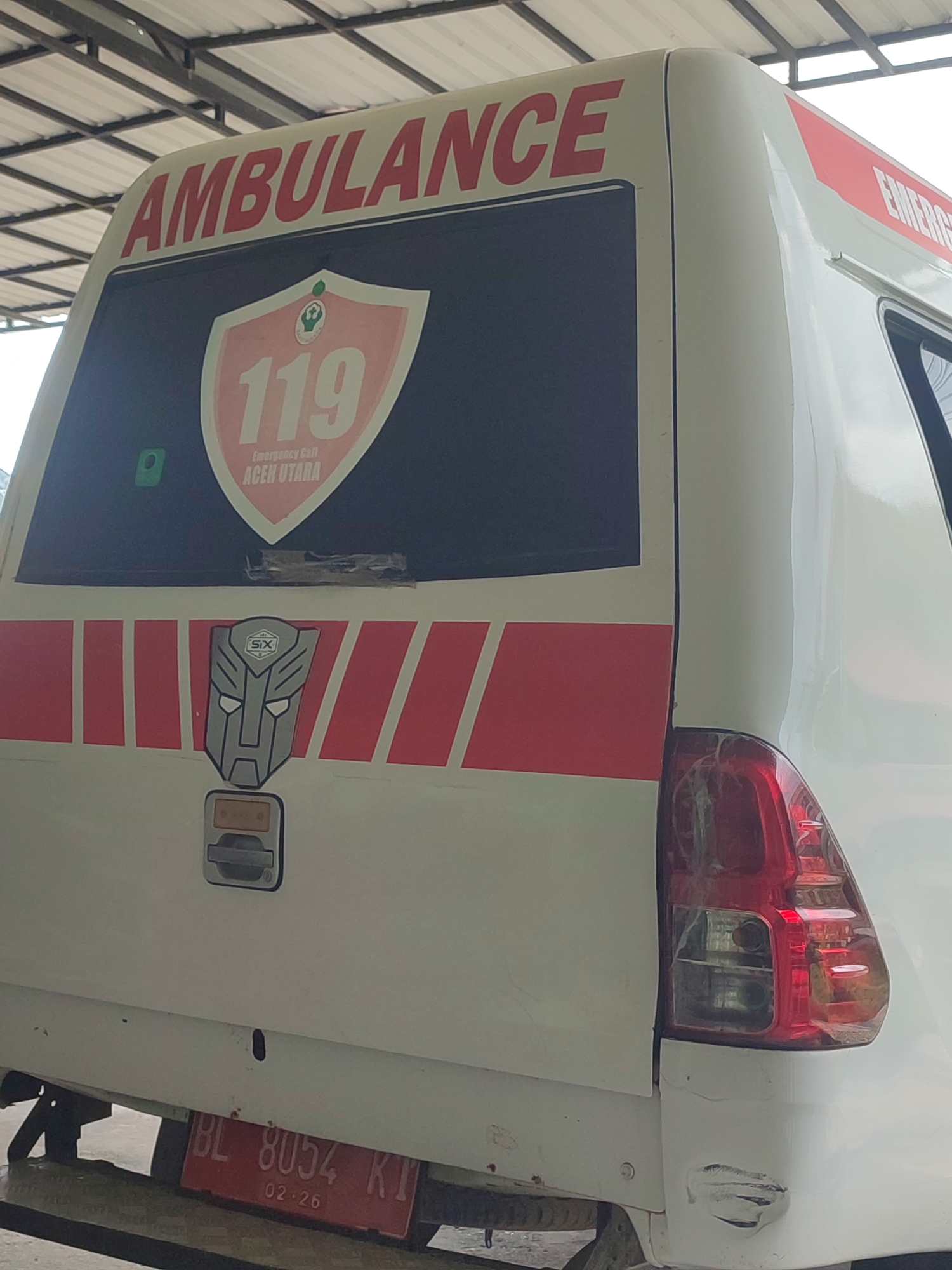 The image shows the rear of a white ambulance parked outdoors under a metal roof structure. The ambulance has "AMBULANCE" written in red letters on its upper rear window. Below this, a shield-shaped emblem with "119" and "Emergency Call ACEH UTARA" is visible. A stylized Transformer logo is also present on the side of the ambulance. The rear lights are red and visible. A red license plate with white text "BL 8054 KI 02-26" is affixed to the lower rear of the vehicle. The environment appears to be a workshop or service area. No people are visible in the image. The image is taken from a medium distance, focusing on the rear of the ambulance.