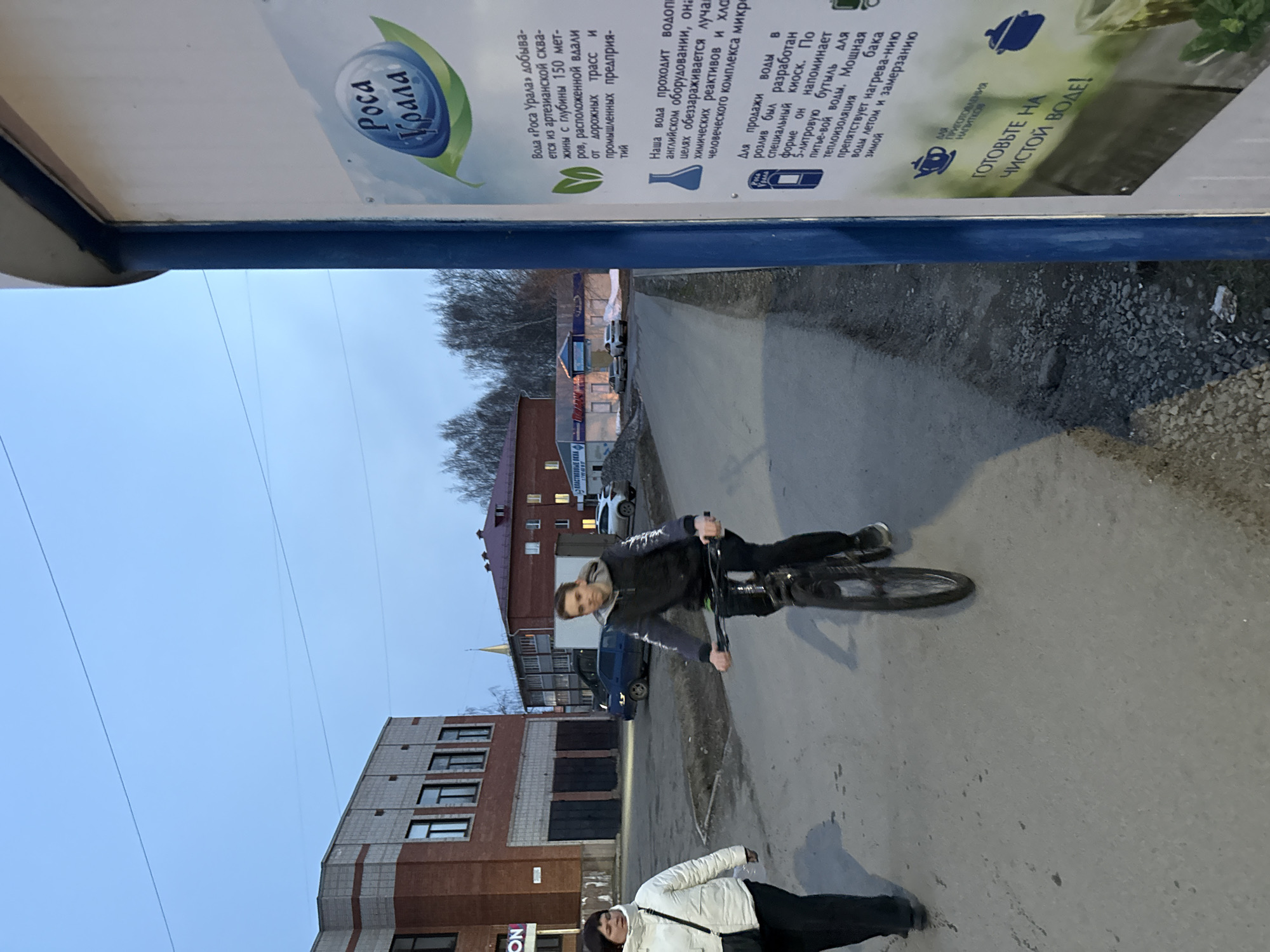 The image depicts a street scene in Votkinsk, Russia. A young man rides a bicycle on a paved road. A woman wearing a light-colored jacket and dark pants walks nearby. A water advertising poster is visible at the top of the image. Buildings are along the street. The sky is overcast.