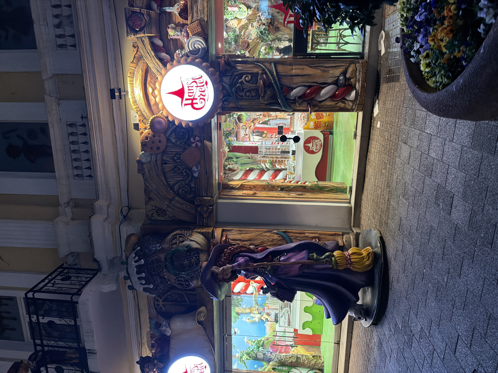 The image shows the exterior of a "Hans & Gretel" shop in Plovdiv, Bulgaria, at night. A statue of a witch stands prominently on the sidewalk in front of the store. The shop's façade is elaborately decorated with elements reminiscent of a fairytale gingerbread house. The storefront is visible with candy cane motifs. A bright sign bearing the "Hans & Gretel" logo is mounted above the entrance. To the right of the store is a planter with flowers. The sidewalk is paved. The architecture of the adjacent building is visible to the left, with a balcony and decorative molding.