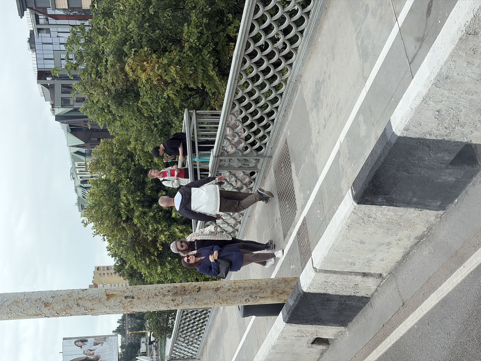 The image depicts a view of a pedestrian bridge in Kaliningrad, Russia, on a cloudy day. A group of four individuals are visible on the bridge. In the foreground, two women walk towards the viewer, one in a dark blue jacket and the other in a plaid shirt. Behind them, two men walk away from the viewer, one in a black jacket and the other in a white jacket with dark sleeves. The bridge has decorative railings and is bordered by concrete barriers. In the background, lush green trees are seen alongside modern buildings with angular roofs and large glass windows. A billboard with images of two women is visible in the far background. The scene appears to be a public walkway, possibly adjacent to a street or waterway.