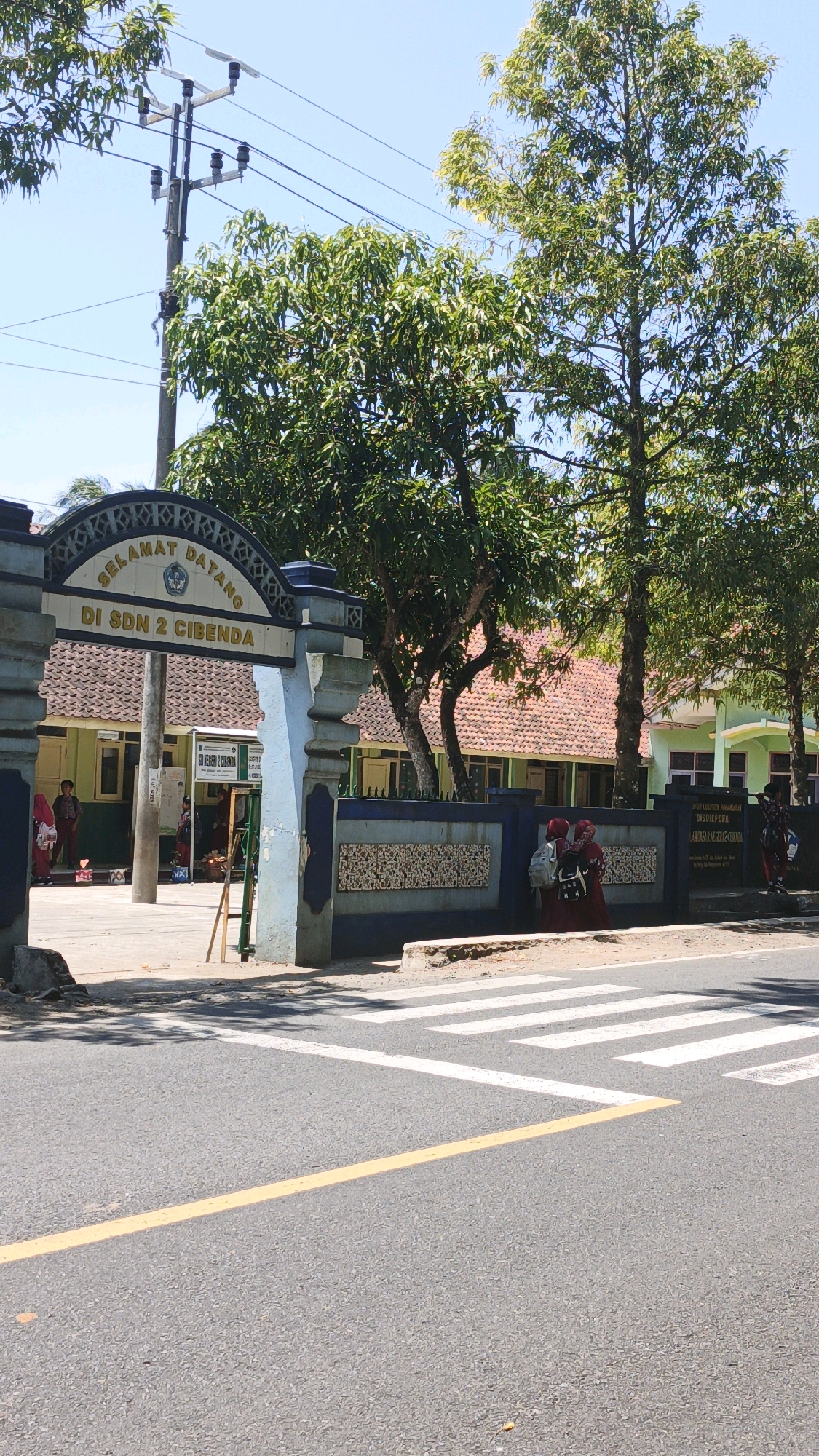 The image shows the entrance to SDN 2 Cibenda, a primary school located in Cibenda, Indonesia. The entrance gate features a blue archway with the words "SELAMAT DATANG DI SDN 2 CIBENDA" (Welcome to SDN 2 Cibenda) inscribed in yellow lettering. Behind the gate, the school building has a red tiled roof and green walls. Several trees with lush green foliage surround the entrance, casting shadows on the pavement. A crosswalk with white stripes is visible on the road in front of the school. Two young students in red uniforms are standing near the entrance, facing the school. Another student, also in a red uniform, is walking away from the school and towards the road. The sky is clear and blue, indicating a sunny day. A utility pole with electrical wires stands to the left of the gate.