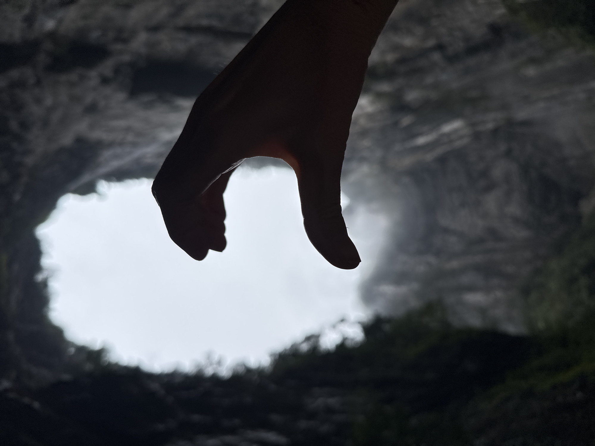 A low-angle shot captures a hand, silhouetted against the backdrop of a karst sinkhole. The fingers are slightly curved, appearing to reach downwards. The sinkhole, known as Xiaozhai Tiankeng, is located in the Wulong District of Chongqing, China. The sinkhole's opening is visible as a large white area, surrounded by the dark, rocky walls of the sinkhole. There is some greenery present along the sides of the sinkhole. The lighting is subdued, giving a stark contrast between the dark foreground and the bright opening.
