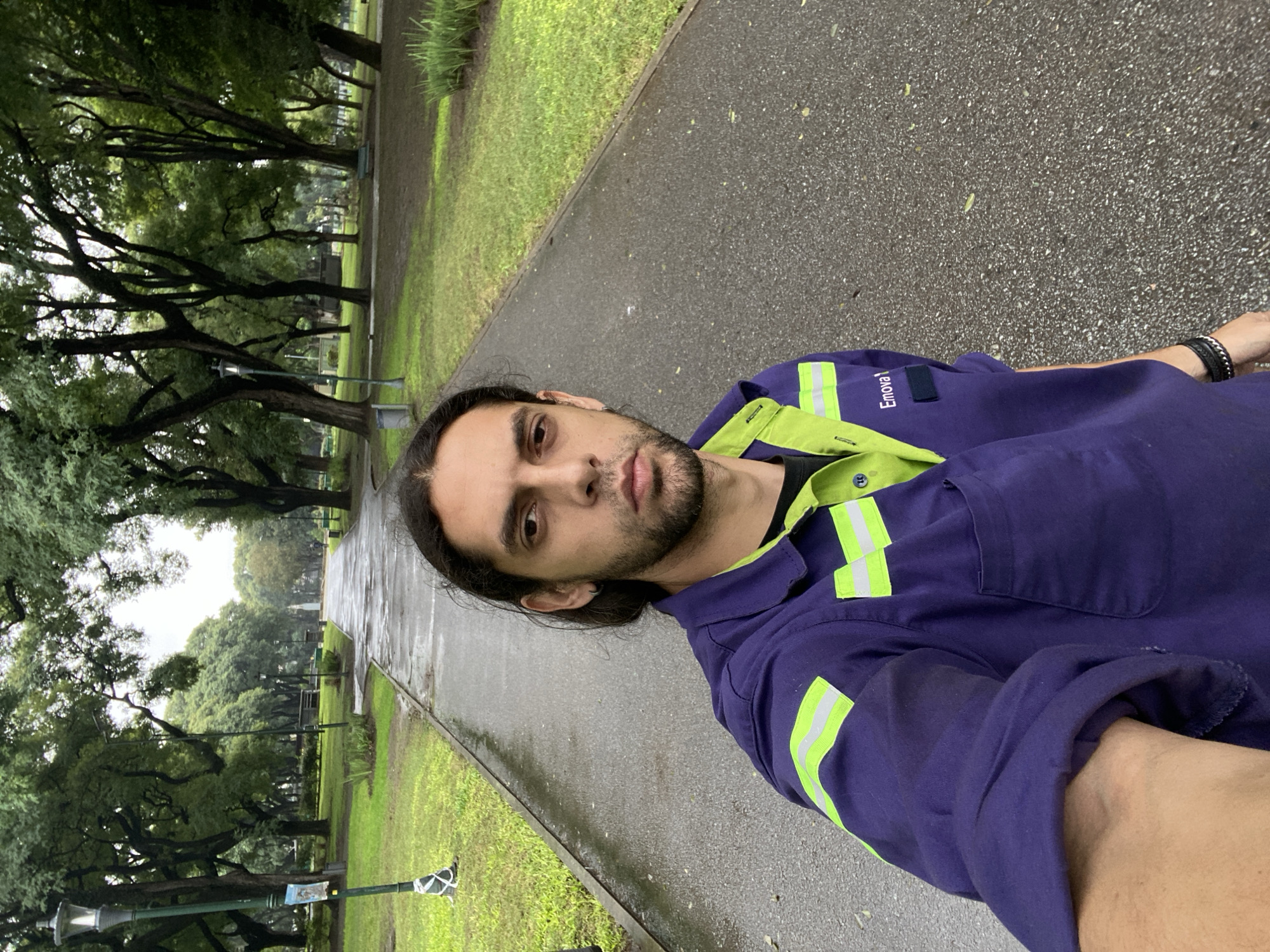 This image is a vertically oriented selfie, originally taken horizontally and rotated 90 degrees counter-clockwise, featuring a man in an outdoor park setting, likely in Buenos Aires, Argentina.
The subject is a man with long dark hair, a beard, and a neutral, serious expression, looking directly at the camera. He is wearing a dark blue work uniform with a light green collar and prominent neon yellow-green reflective stripes on both shoulders and upper arms. A black t-shirt is visible beneath the uniform. On the right chest area of his uniform, the word "Enova!" is clearly visible as a white logo. He wears a black, possibly braided, bracelet on his left wrist.
He is positioned on a paved path, which appears wet, indicating recent rain or damp conditions. Adjacent to the path is a strip of wet green grass, bordered by numerous large trees with dark trunks and dense green foliage, creating a shaded environment. In the background, more trees, additional paths, and what appears to be a streetlamp are visible, consistent with a public park. The sky is overcast, casting a soft, diffused light over the scene. The overall impression is a damp, perhaps cool, daytime setting in an urban green space.